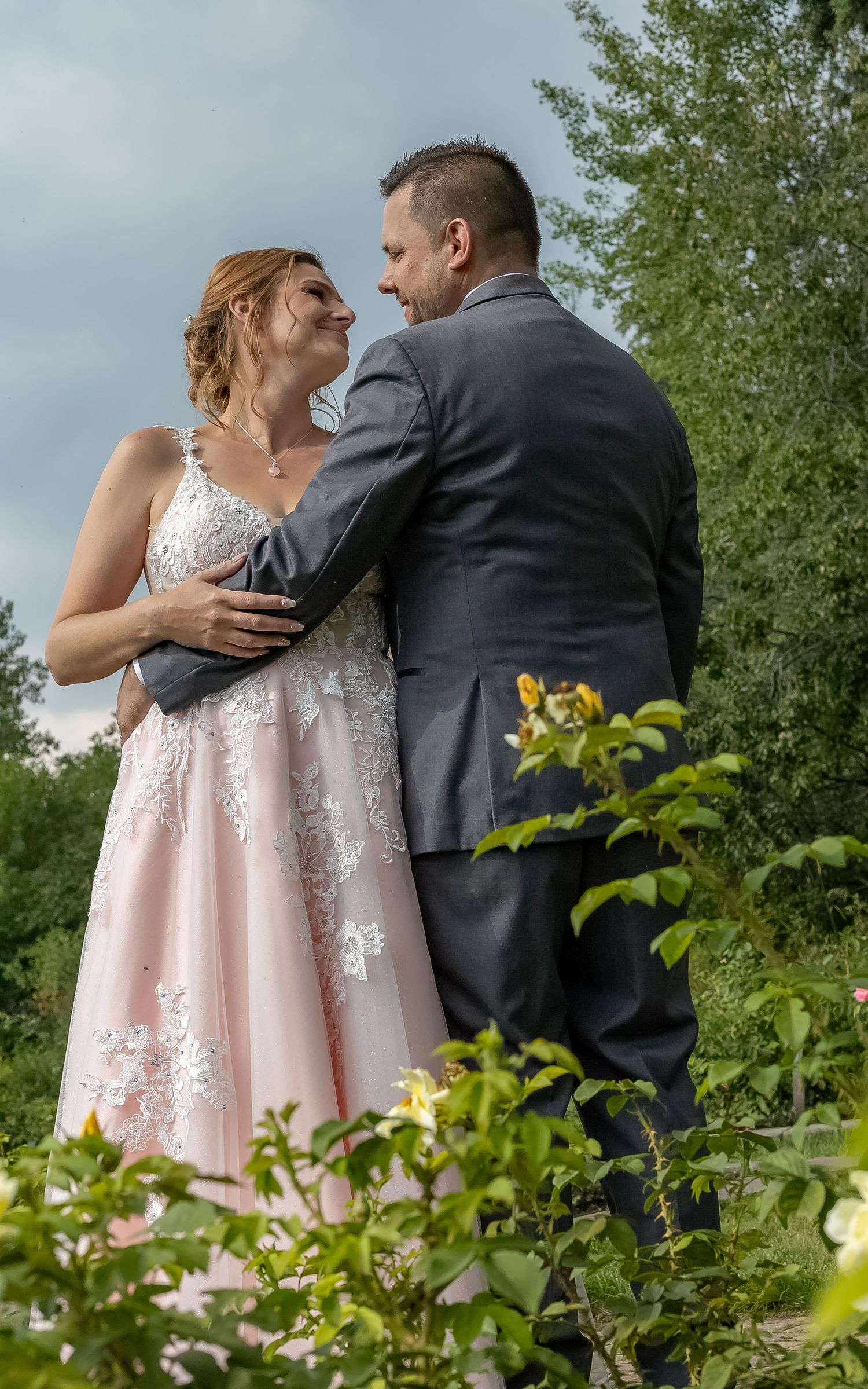 Bride in pink lace gown embraces groom in gray suit among garden greenery.
