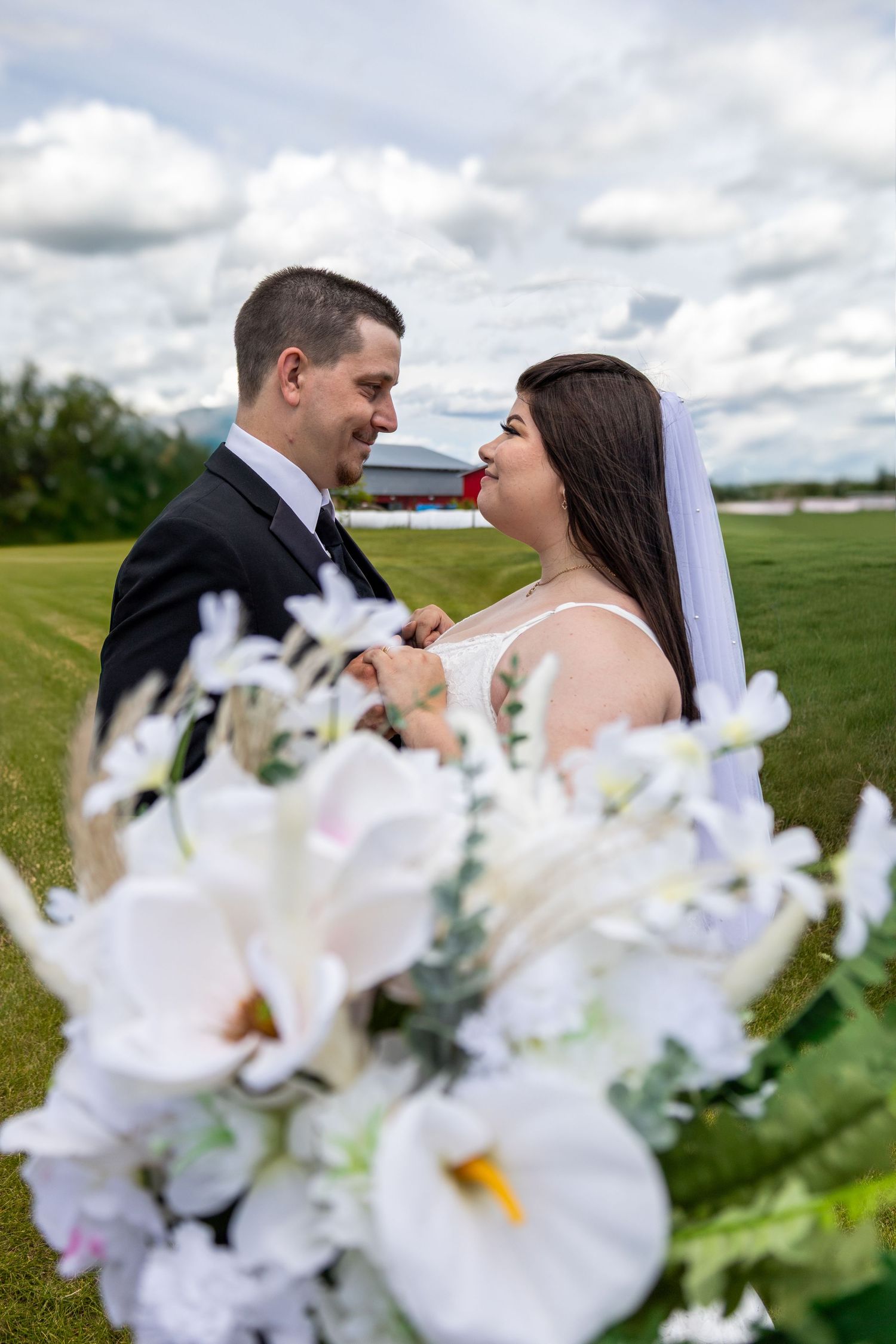 Bride and groom share romantic moment in green field with white flower bouquet in foreground.