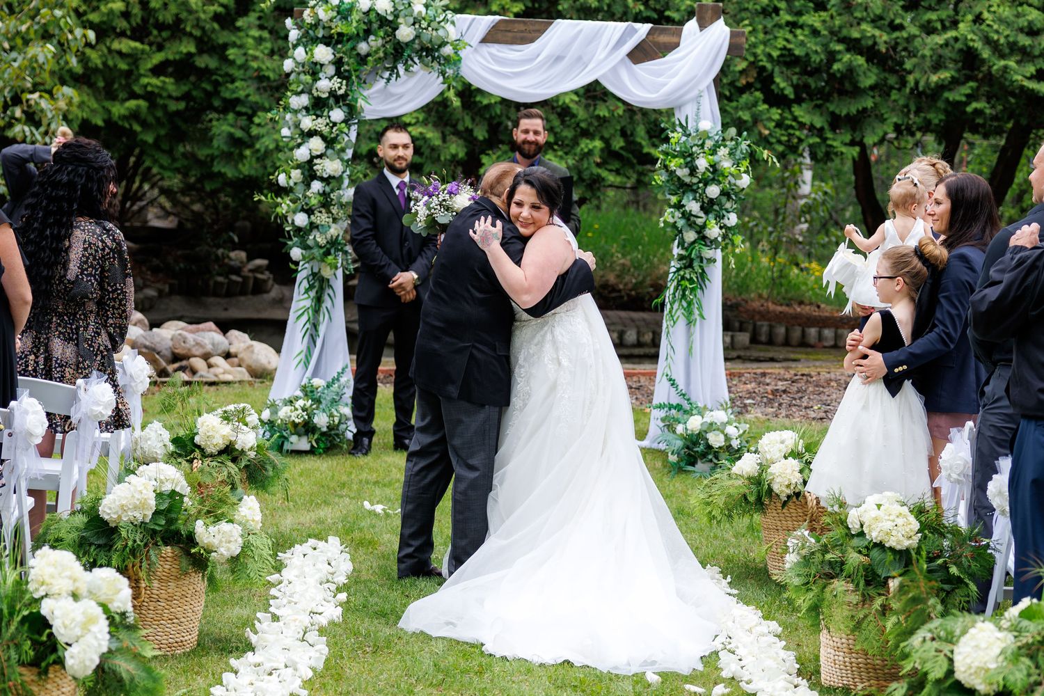 Bride and groom share first kiss under a floral arch surrounded by white hydrangeas at outdoor ceremony.