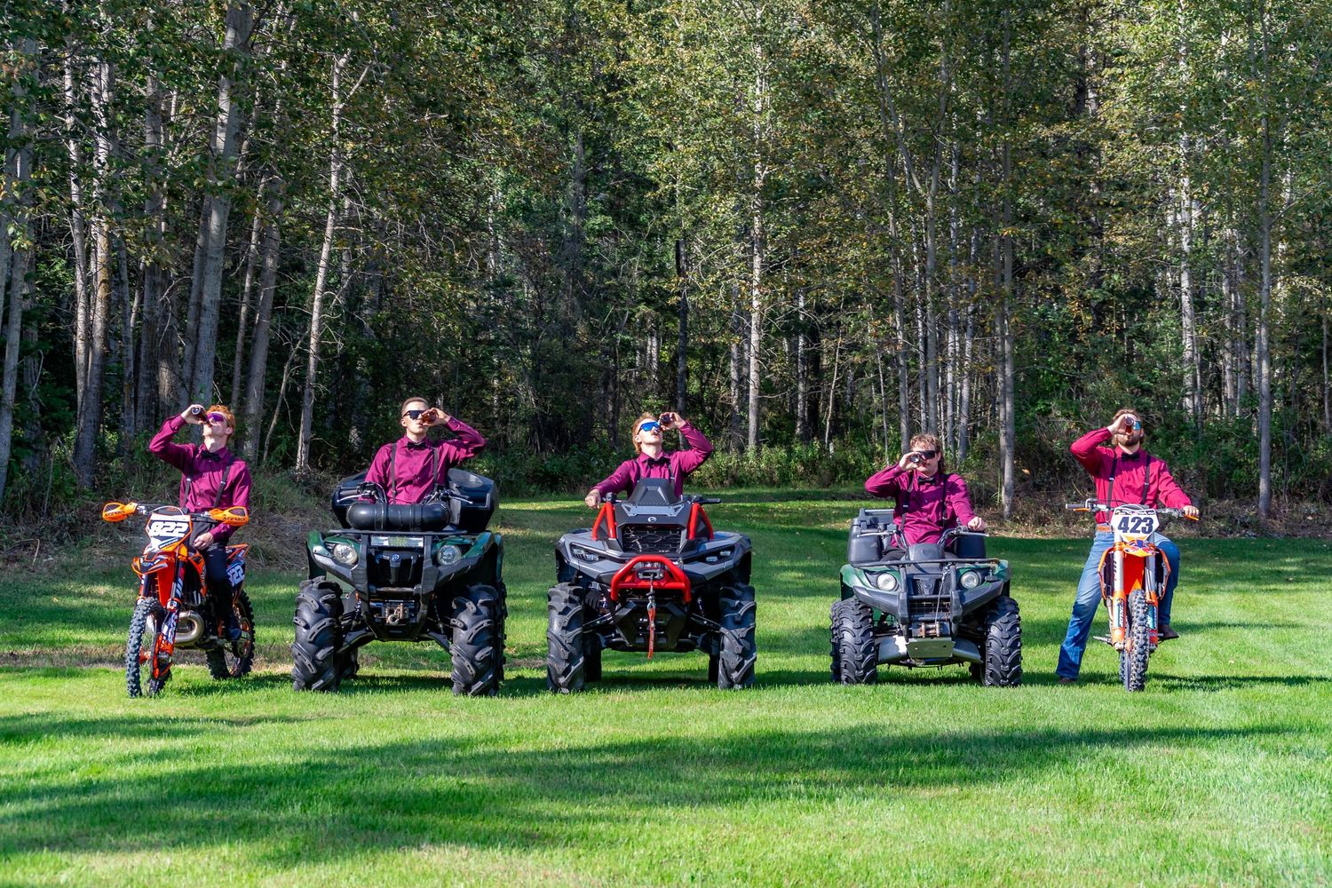 Groomsmen in burgundy shirts posing on ATVs and dirt bikes in green field.