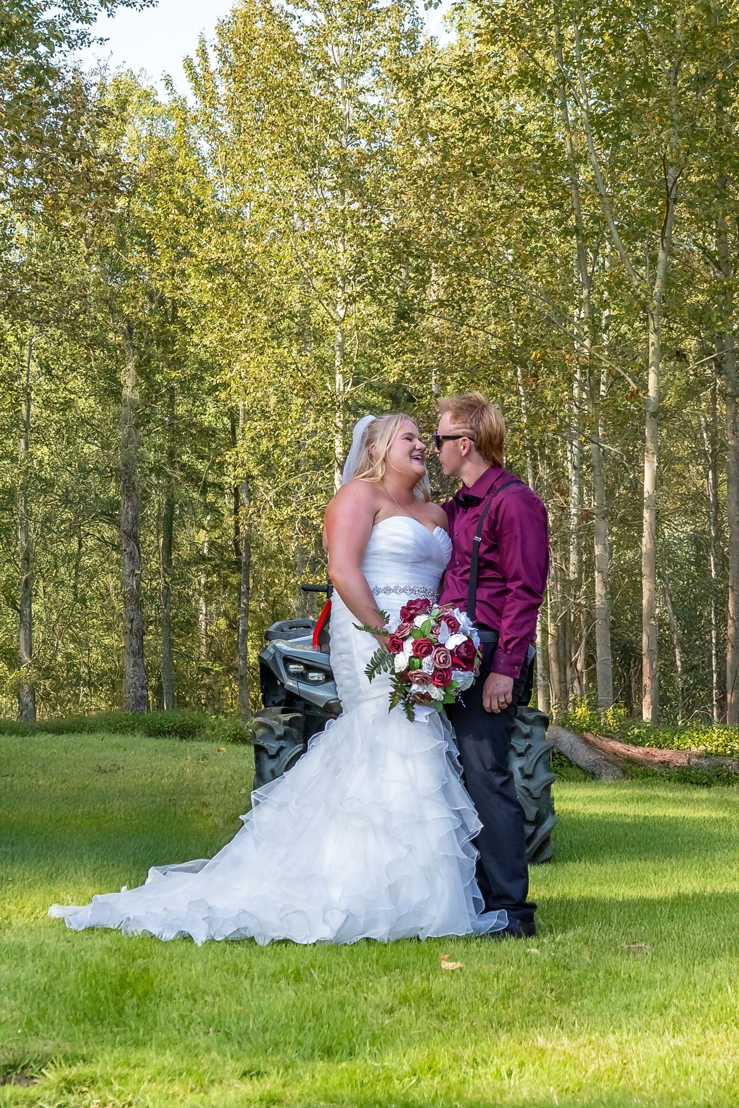 Bride in white ruffled gown and groom in burgundy shirt sharing romantic moment outdoors.