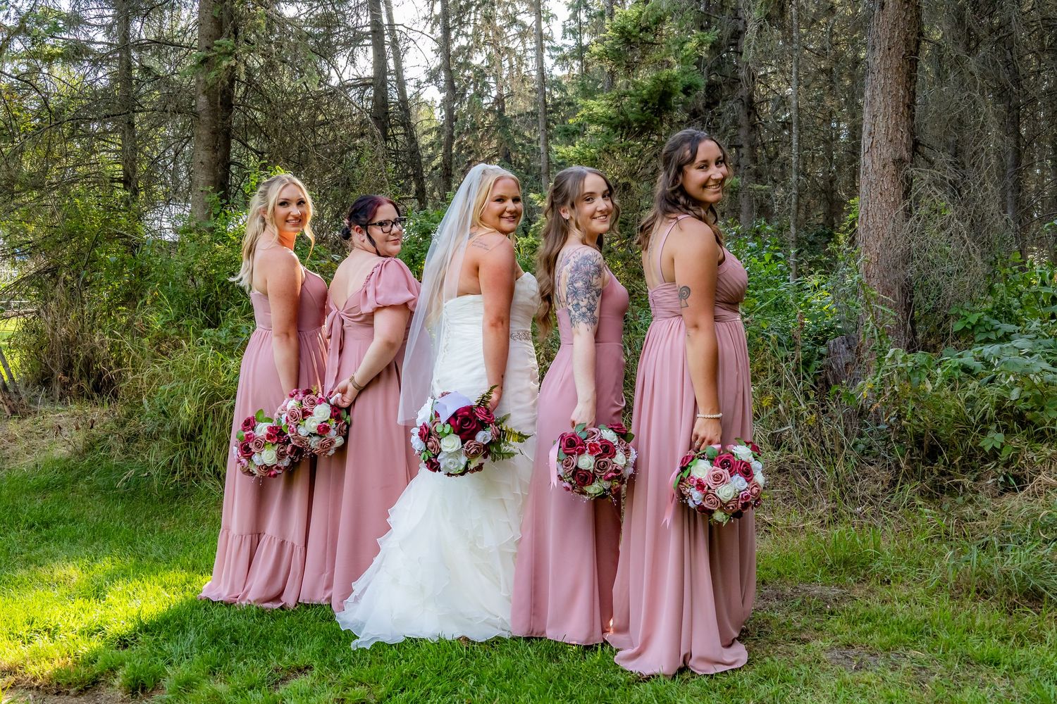 Bride and bridesmaids in pink dresses posing with bouquets in wooded outdoor setting.