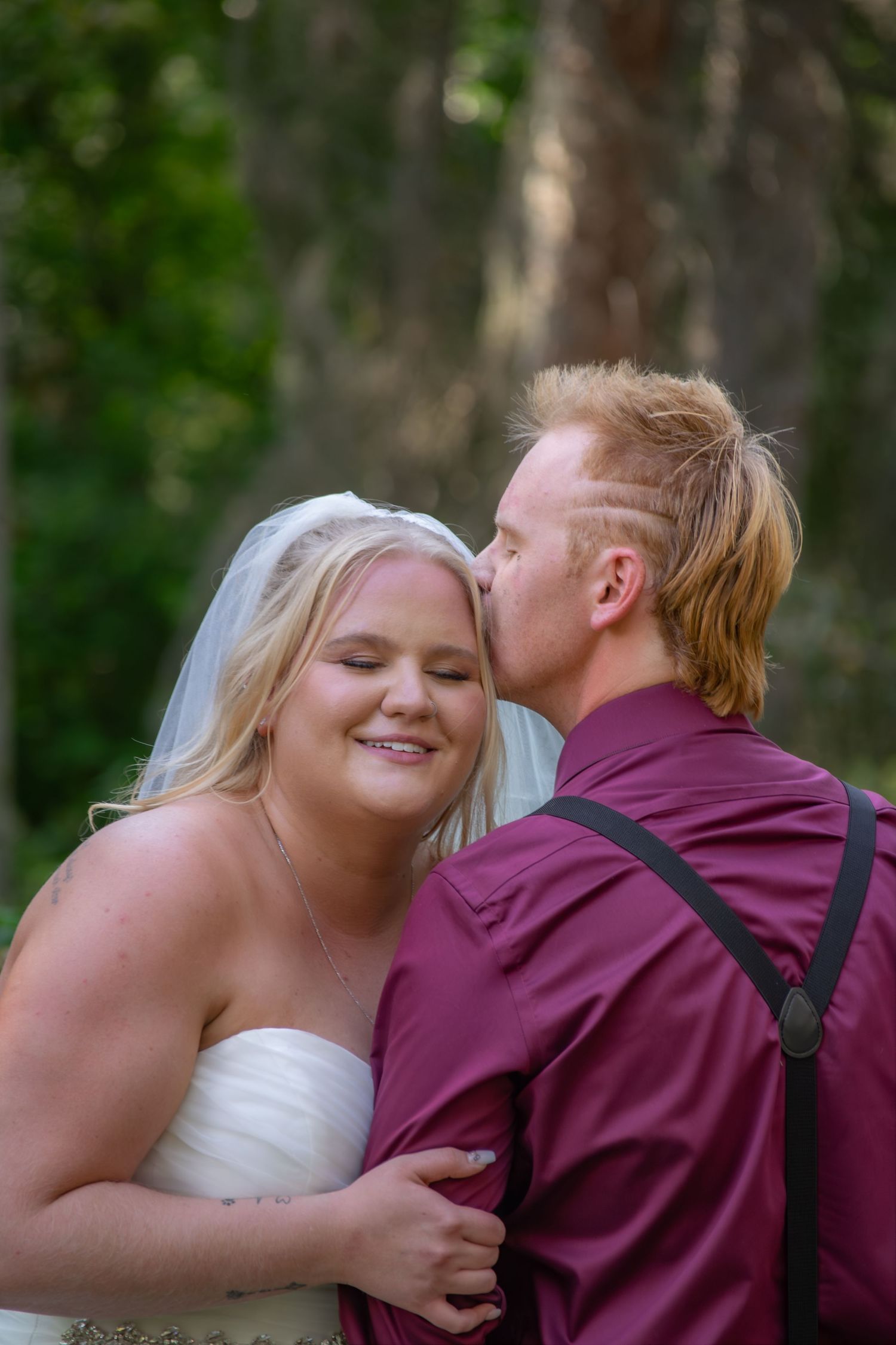 Groom in burgundy shirt kissing smiling bride on the cheek in a romantic forest setting.