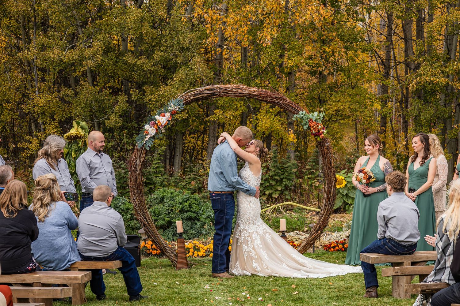 Couple shares first kiss under rustic circular wooden arch adorned with autumn flowers at outdoor fall wedding ceremony.
