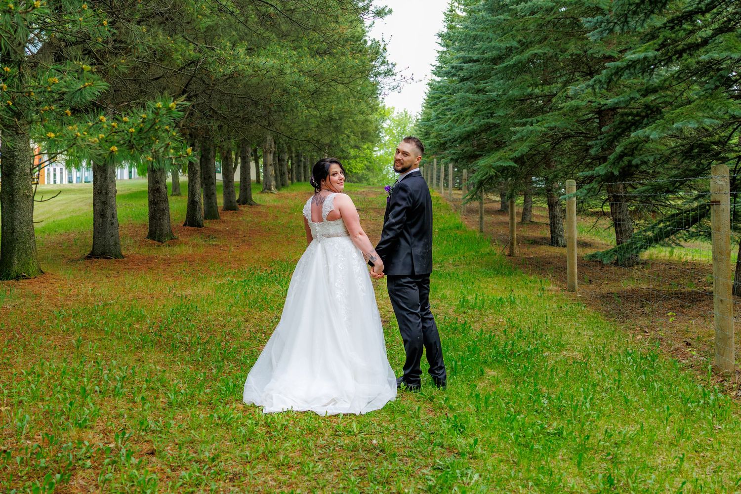 Bride in white ball gown and groom hold hands walking between tall evergreen trees in park.