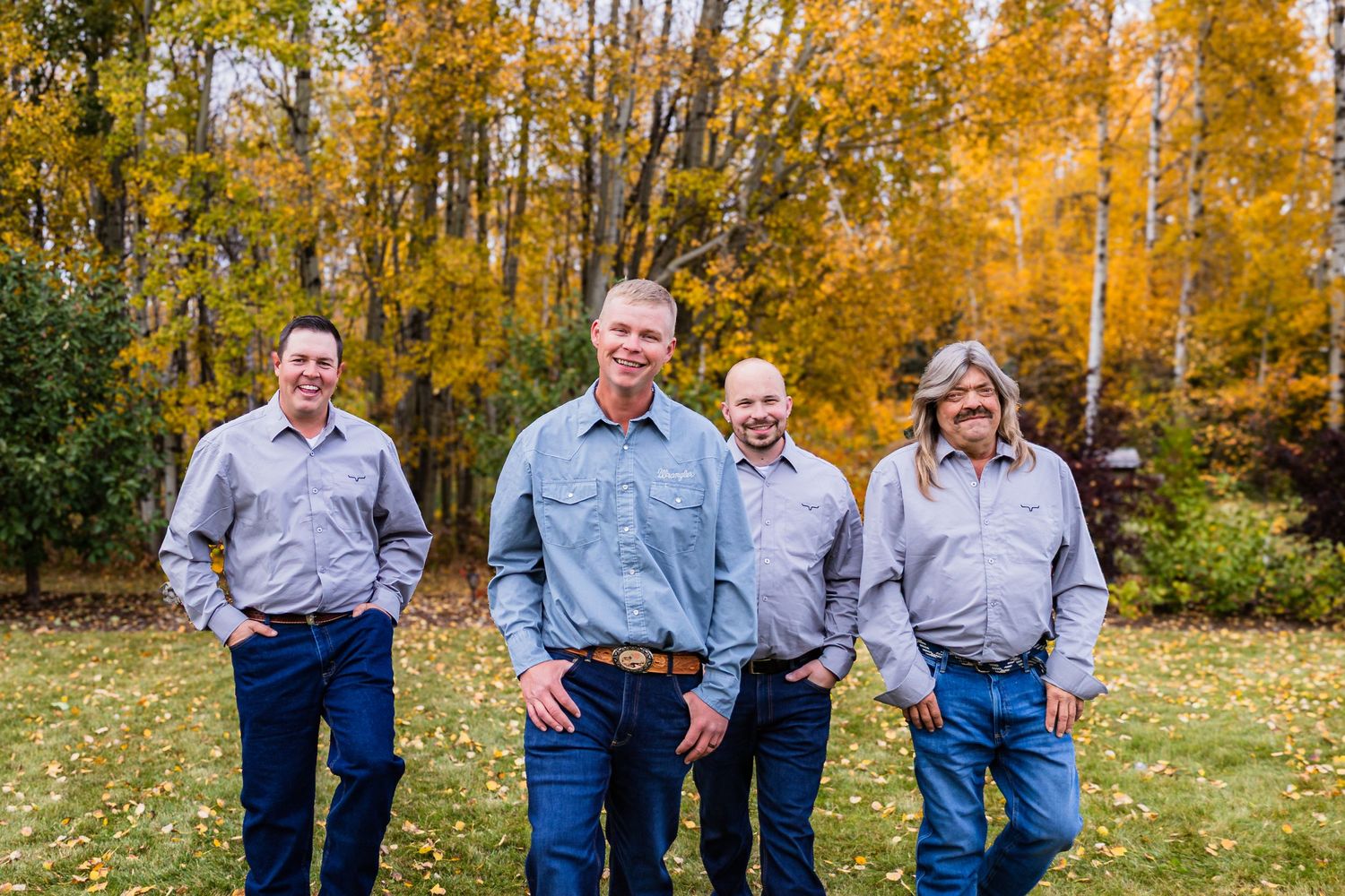 Four groomsmen in gray shirts smiling while walking through golden autumn foliage outdoors.