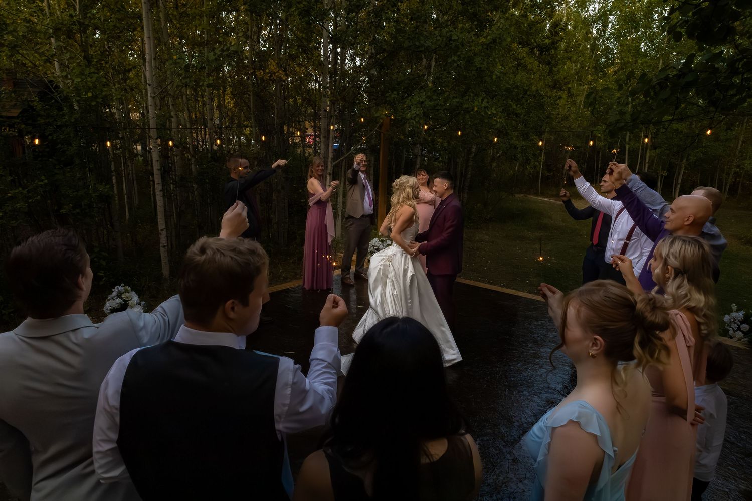 Bride and groom sharing first dance surrounded by guests under string lights in a wooded outdoor venue at dusk.