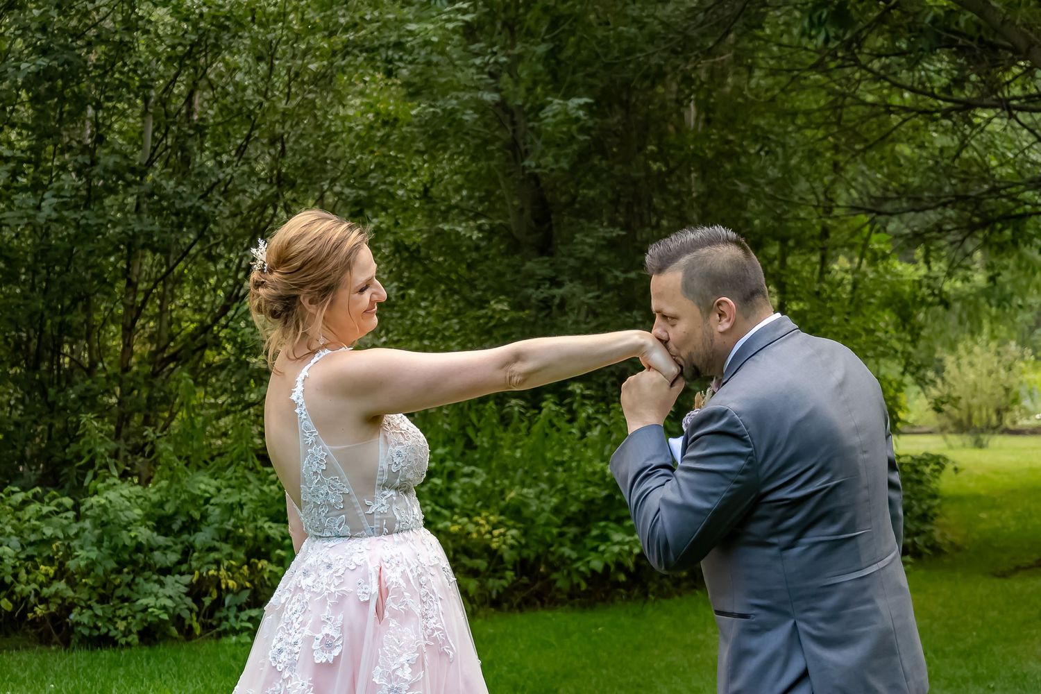Groom in grey suit kisses bride's hand as she extends her arm in a lush green outdoor garden setting.