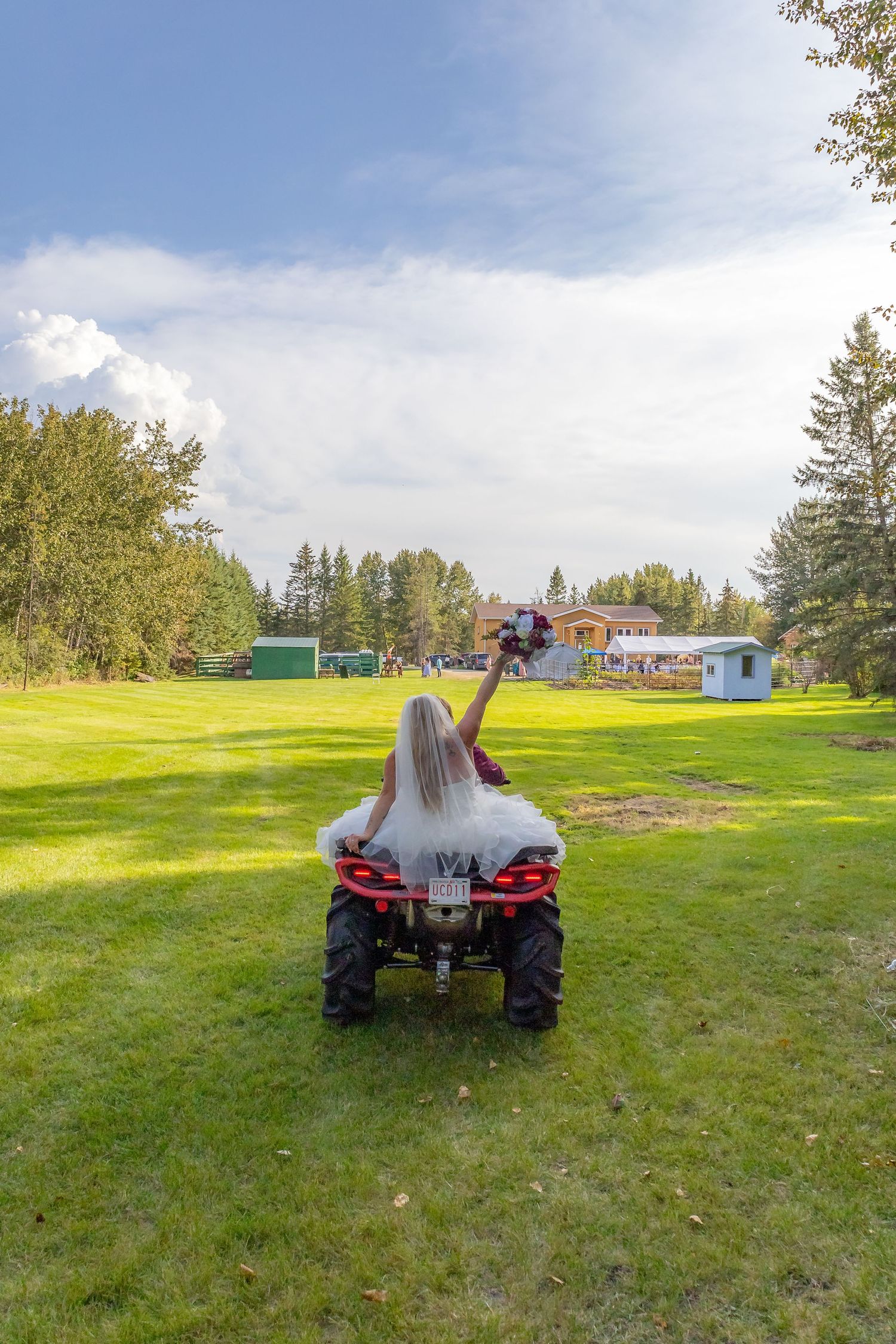 Bride in white wedding dress riding an ATV across a green lawn while taking a selfie.