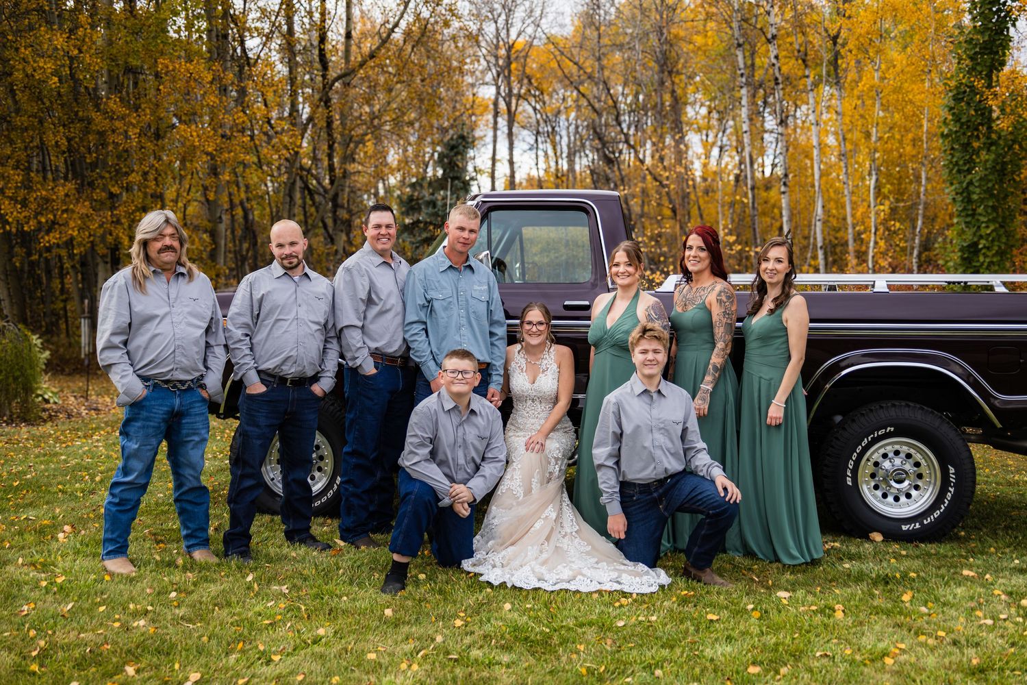 Wedding party posing in front of a vintage dark truck amid autumn foliage, bridesmaids in sage green gowns.