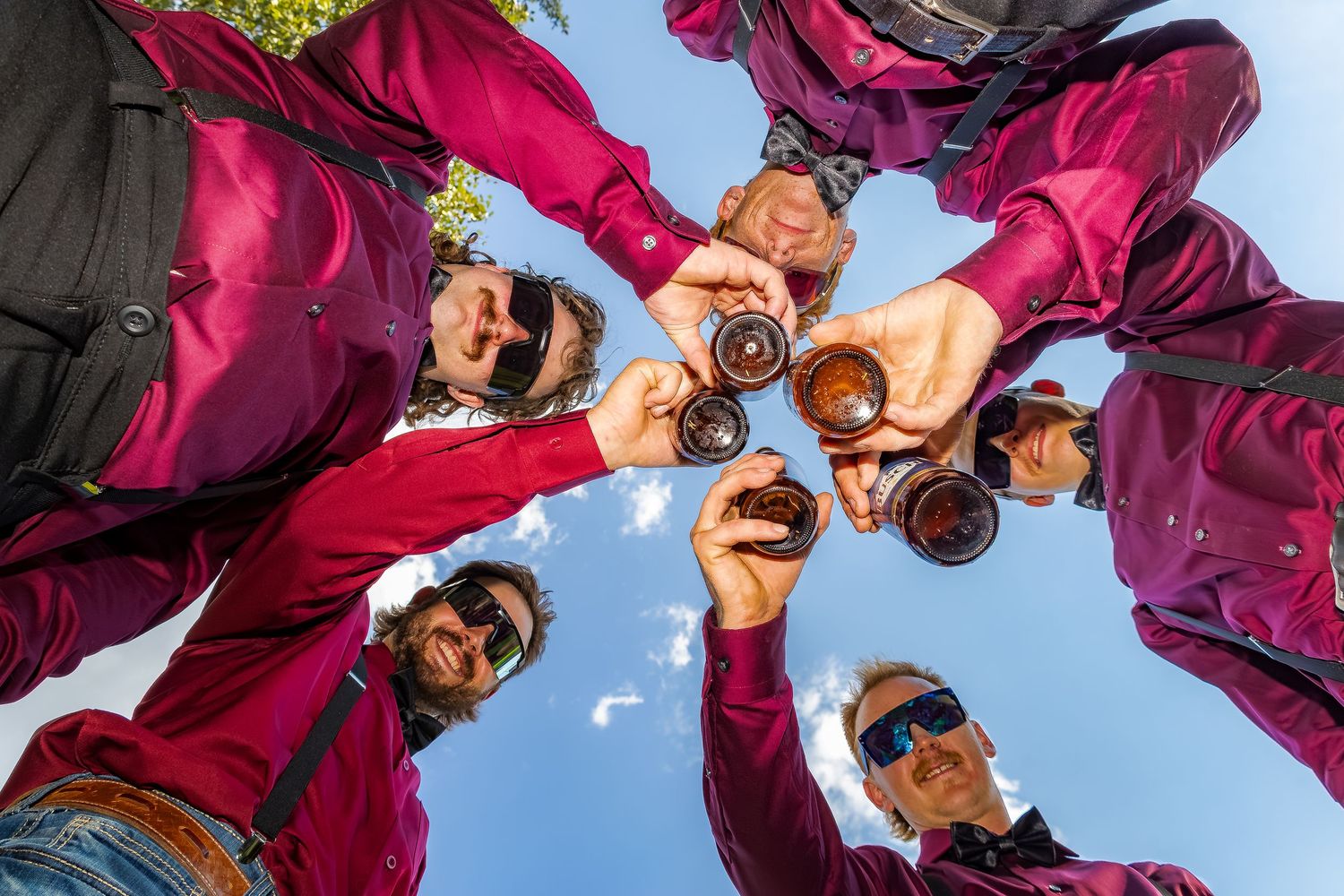 Groomsmen in magenta shirts toast with drinks in a circle captured from below against blue sky.