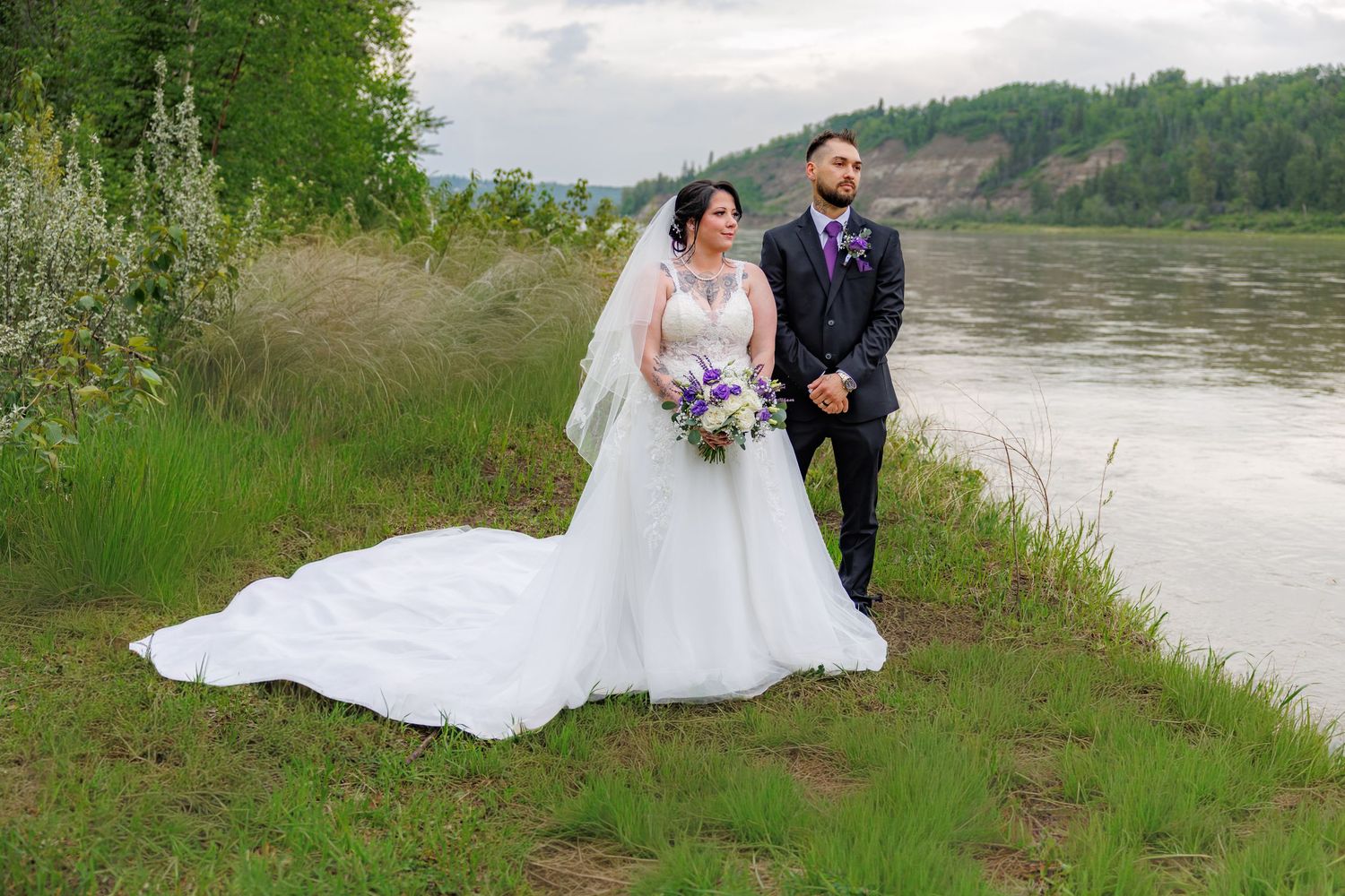 Bride in white wedding gown and groom in dark suit pose by a river with lush greenery on their wedding day.