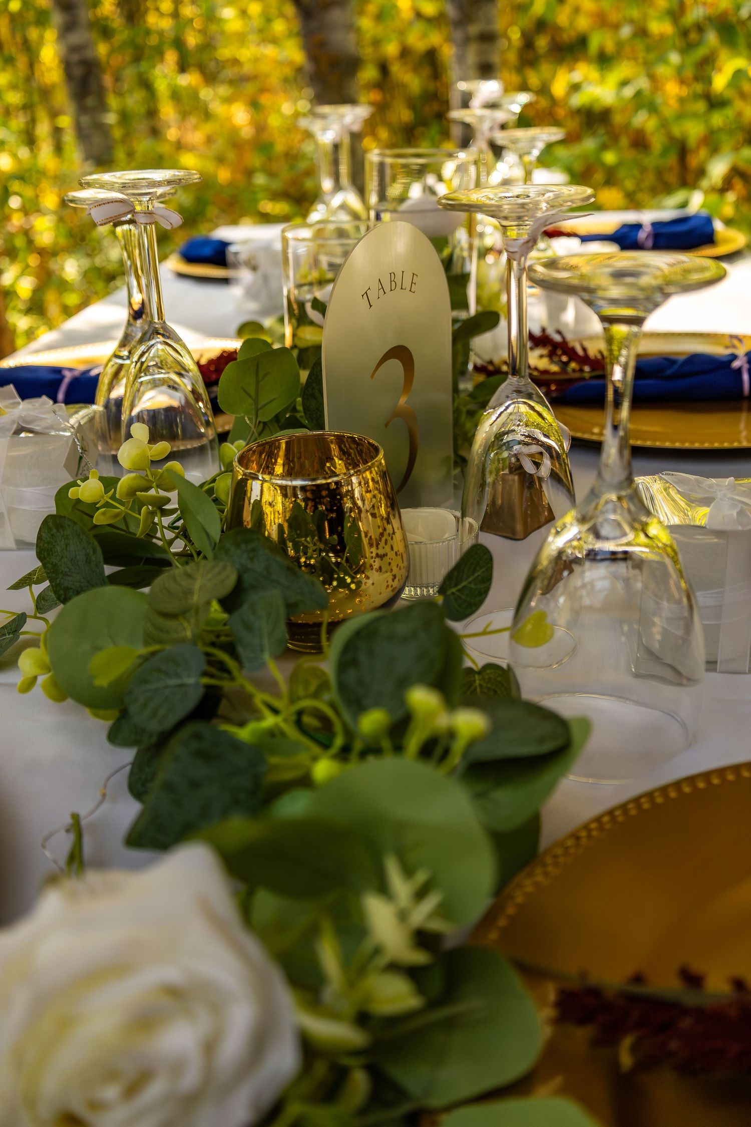 Elegant outdoor wedding reception table set with gold chargers, greenery garland, and gold mercury glass votives.
