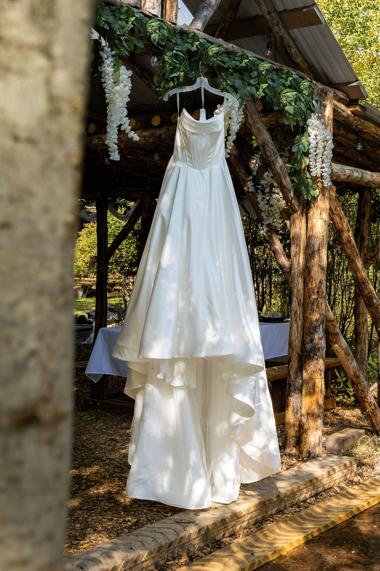 Elegant white wedding dress hanging on a rustic wooden arbor decorated with white wisteria flowers outdoors.