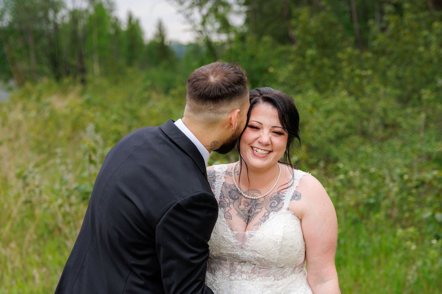 Groom kissing bride on cheek as she smiles during outdoor wedding portrait in lush green setting.