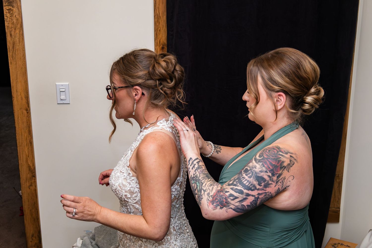 Bridesmaid in green dress helping bride with lace wedding gown before ceremony indoors.