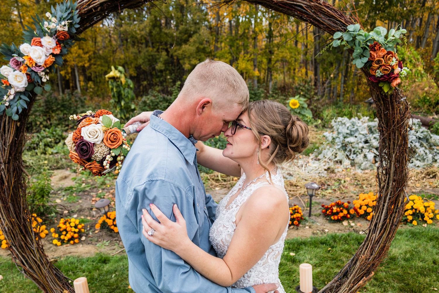 Bride and groom sharing a romantic moment under a rustic floral arch with autumn flowers and greenery.
