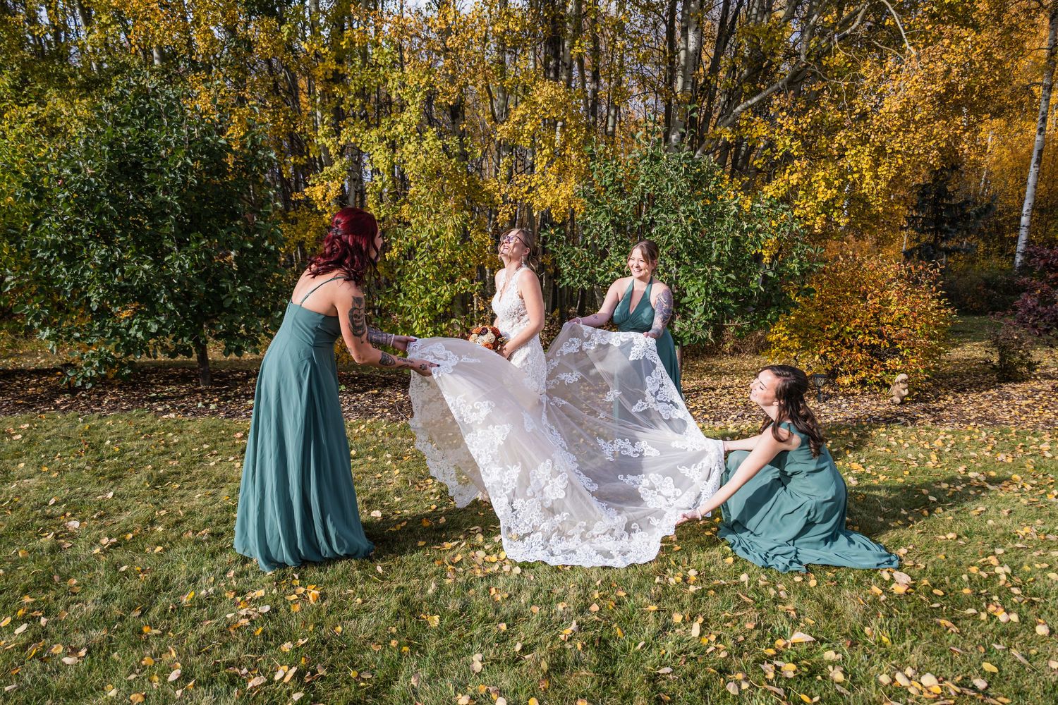 Three bridesmaids in teal dresses fluffing bride's lace wedding gown train outdoors in fall.