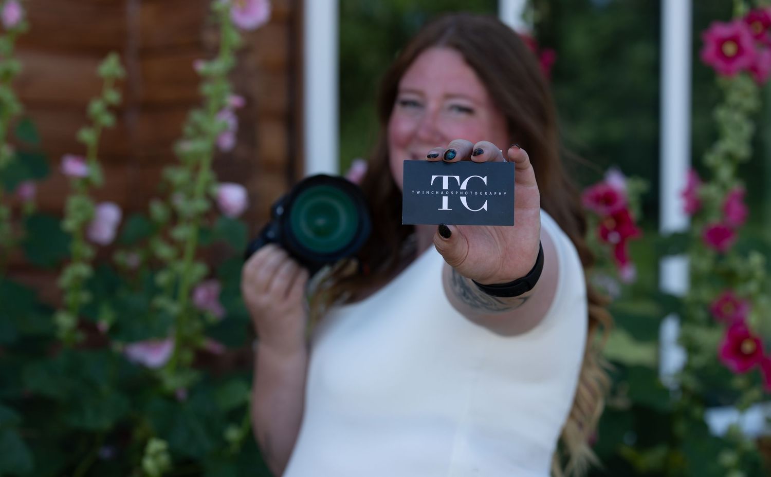 Woman photographer holding camera and TC business card outdoors among pink hollyhocks flowers.