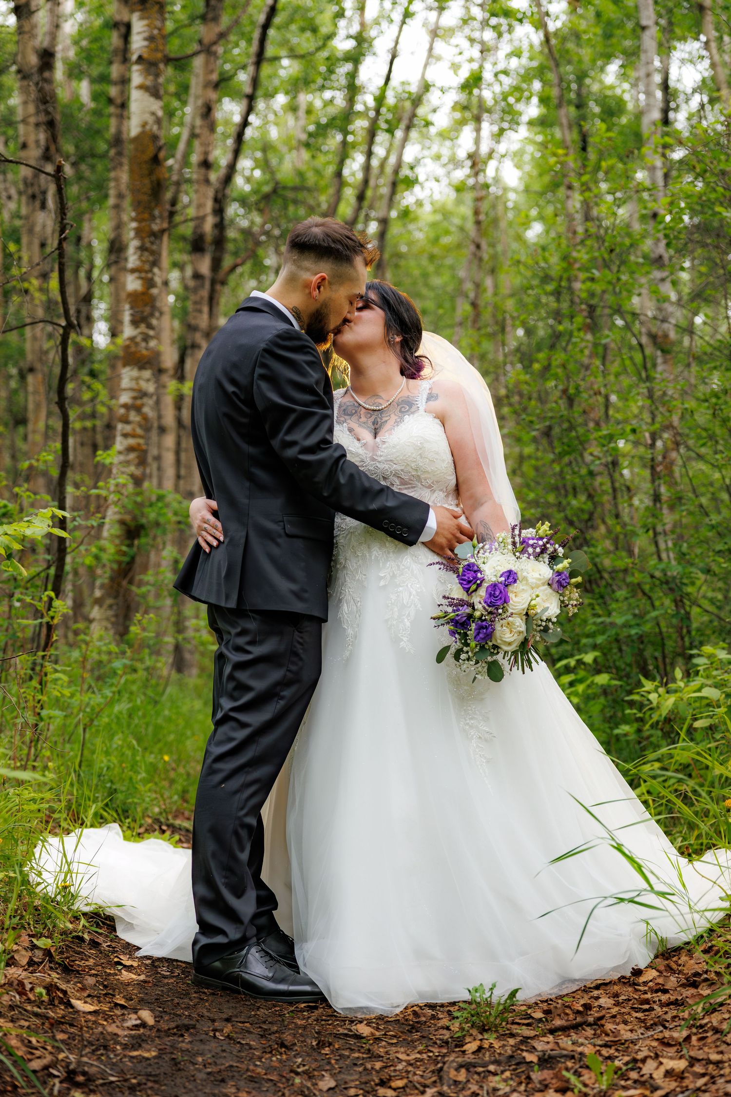 Bride and groom sharing a kiss in a lush green birch forest, bride holding purple and white bouquet.