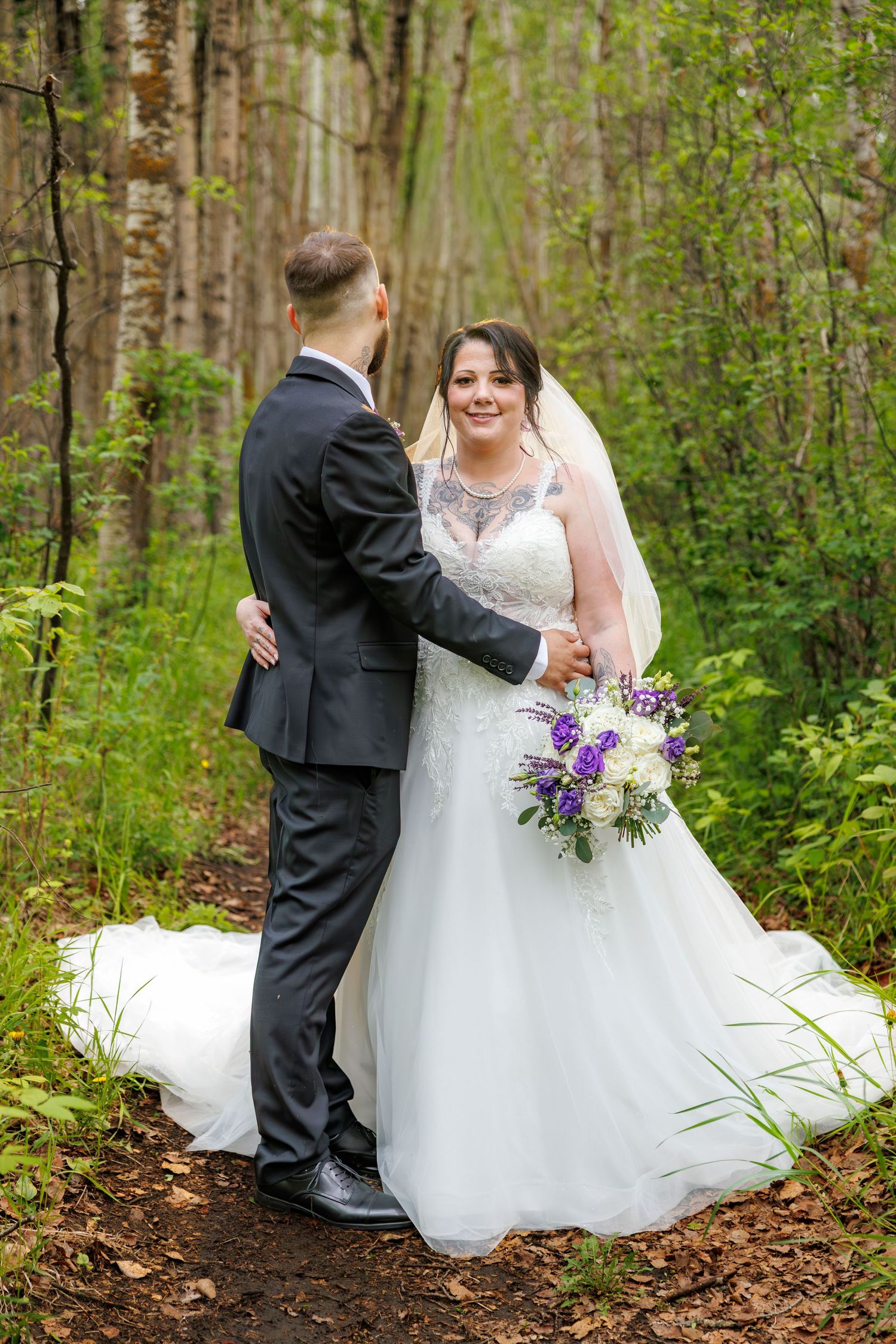 Bride in lace wedding gown holds purple bouquet while embracing groom on a woodland forest path.