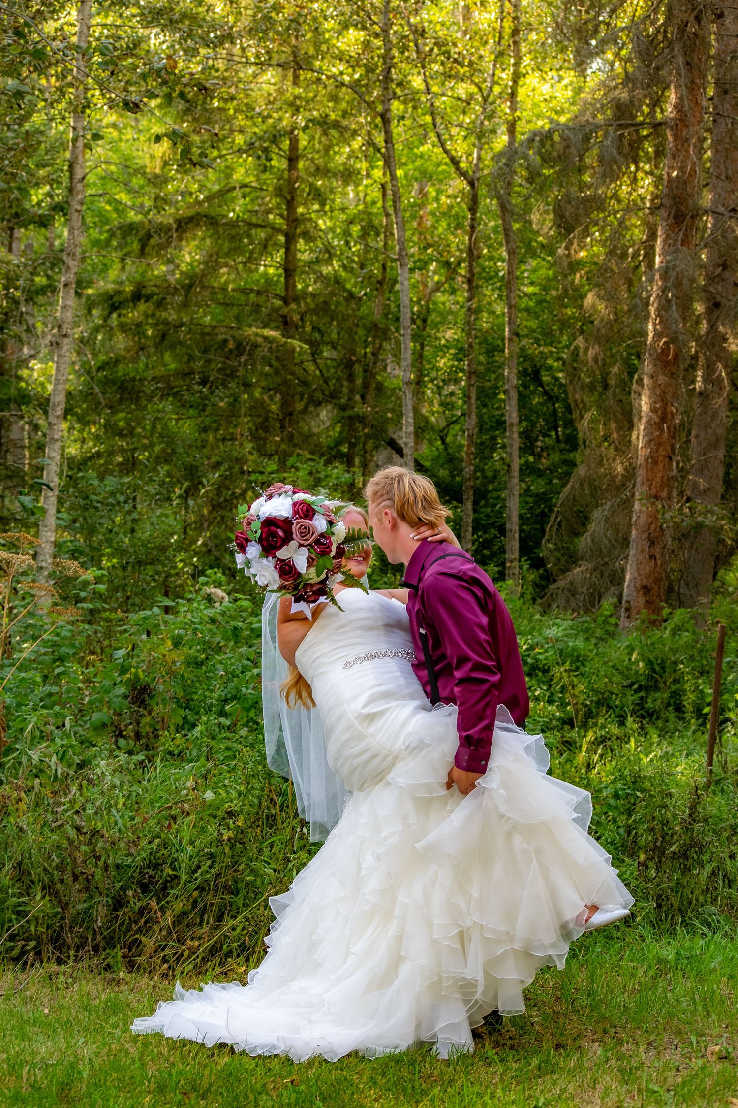Groom in burgundy suit dipping bride in white wedding dress in lush green forest.
