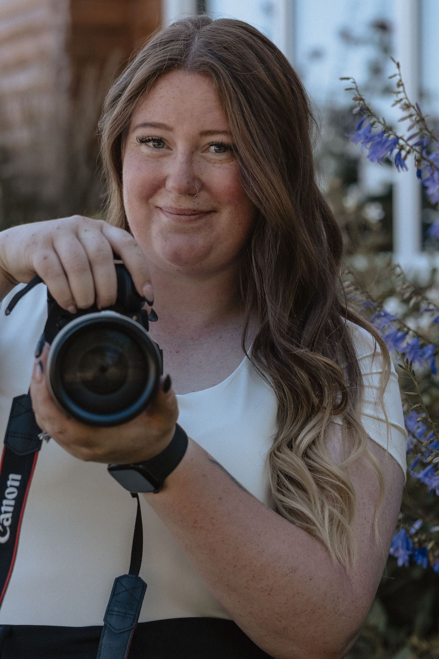 Woman smiling and holding a Canon camera outdoors with purple flowers in the background.