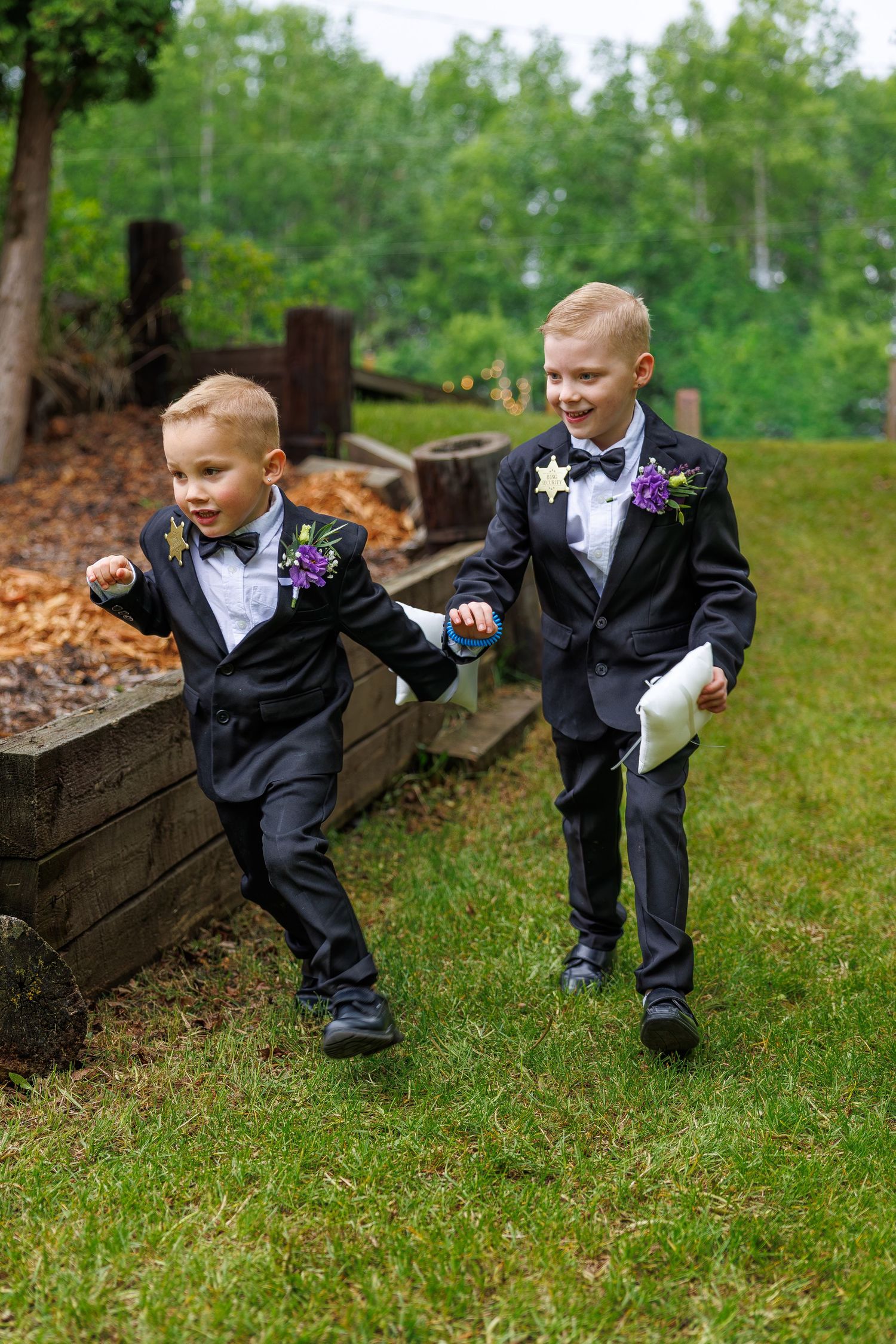Two young ring bearers in matching black tuxedos with purple boutonnieres running on green grass outdoors.