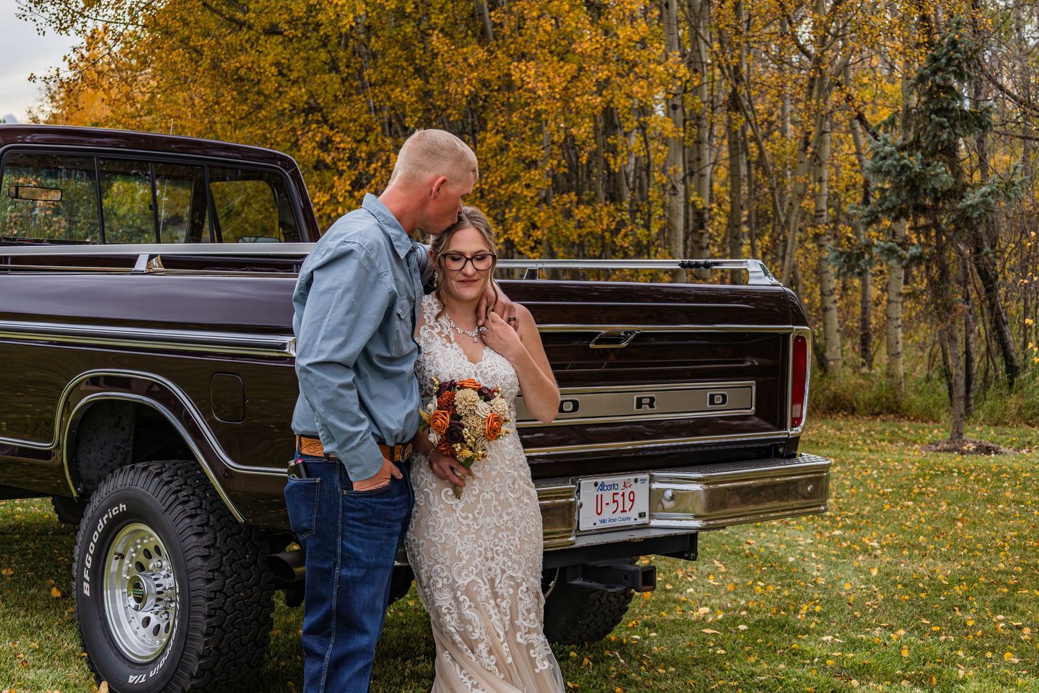Bride in lace gown and groom in denim shirt posing together beside a vintage Ford truck in fall setting.