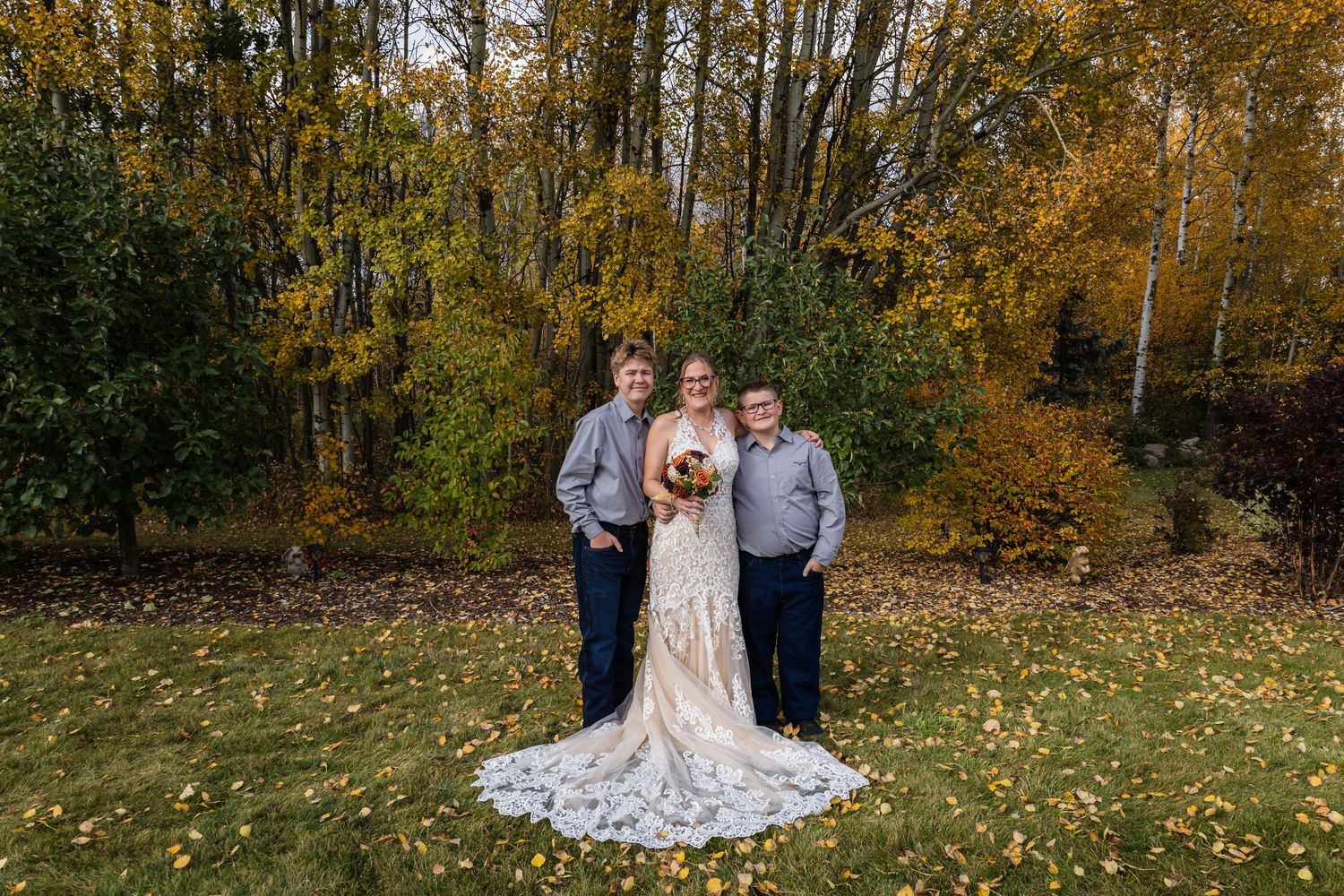Bride in lace gown with bouquet poses with two men in gray shirts amid fall foliage.