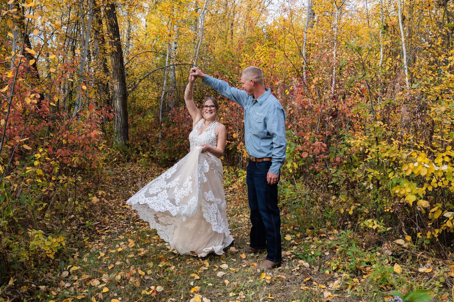 Groom twirling bride in lace wedding dress along a colorful autumn forest path surrounded by fall foliage.