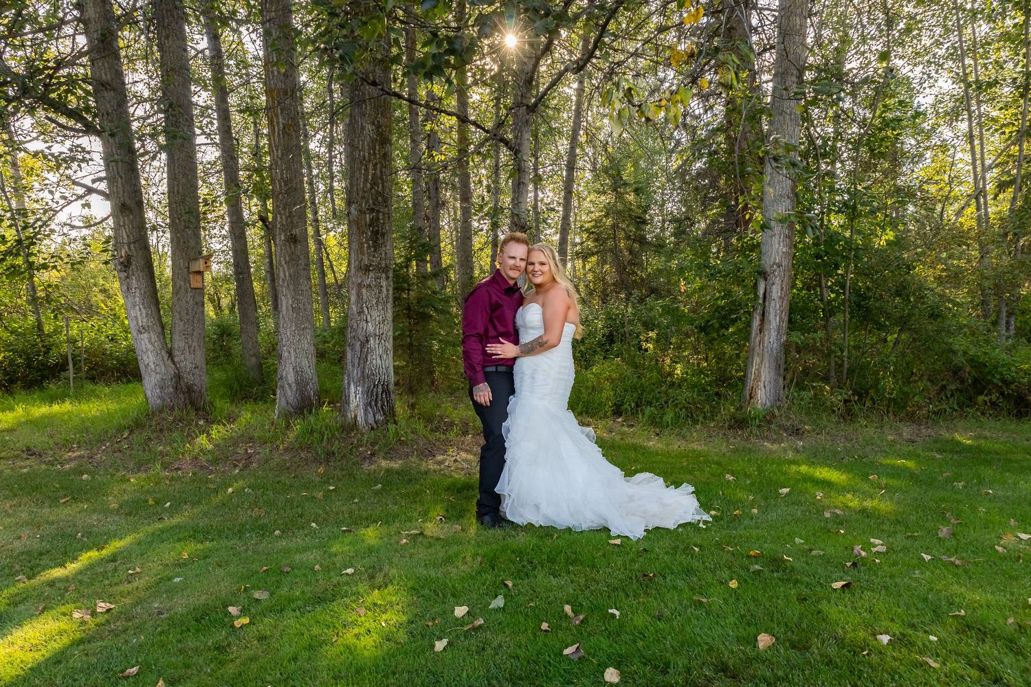 Smiling bride and groom posing together among tall trees in a sunlit outdoor wedding setting.