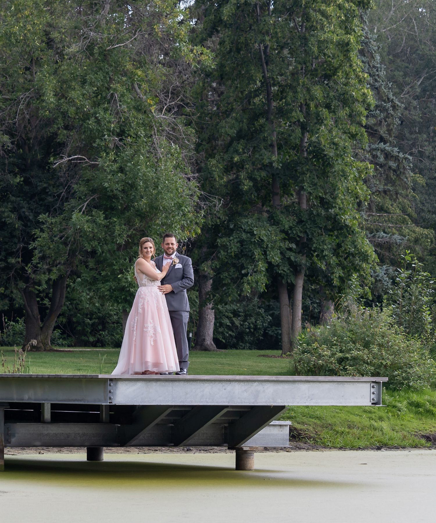 Bride and groom pose on a dock over a pond surrounded by lush green trees.