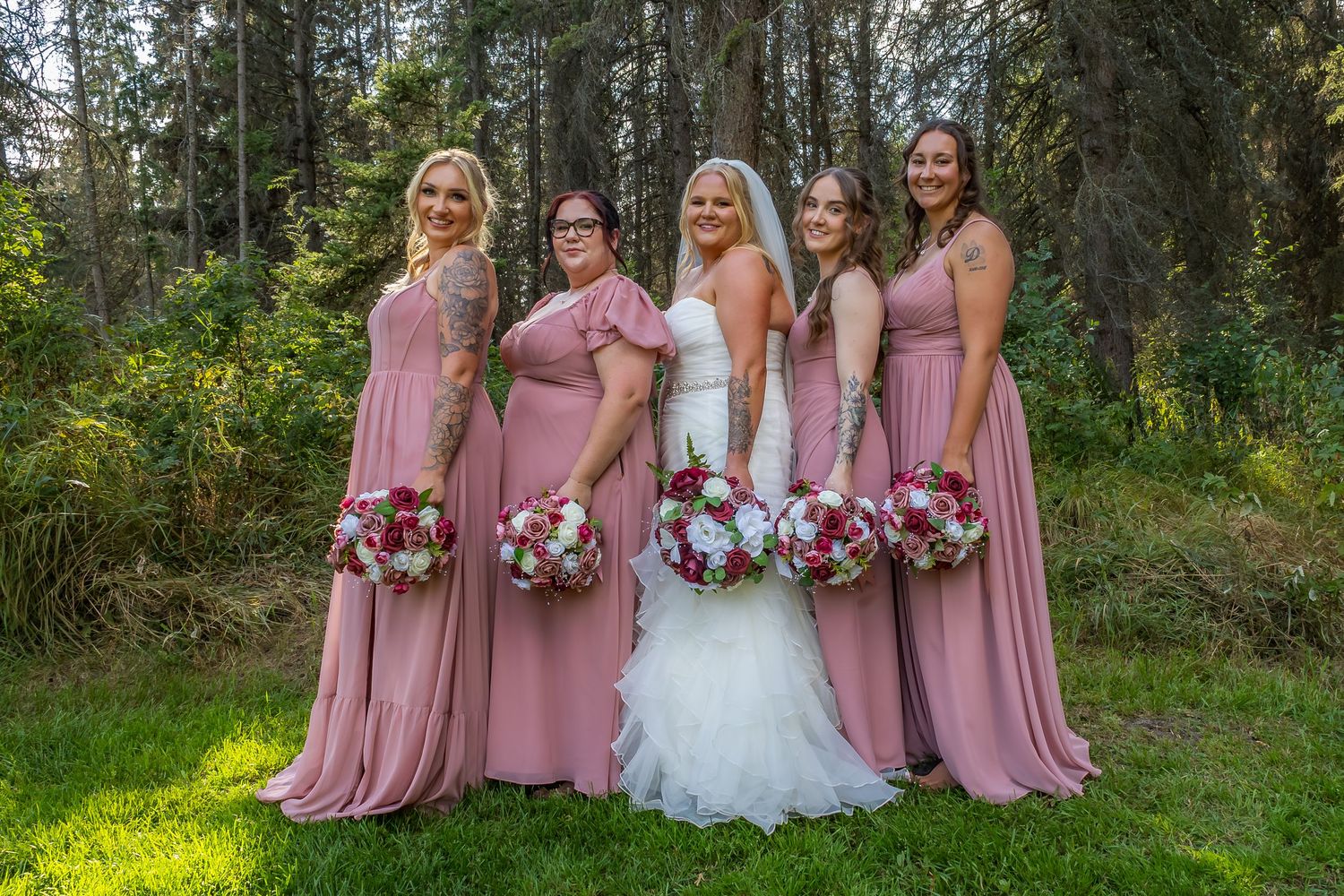 Bride with four bridesmaids in dusty rose gowns holding bouquets in forest setting.