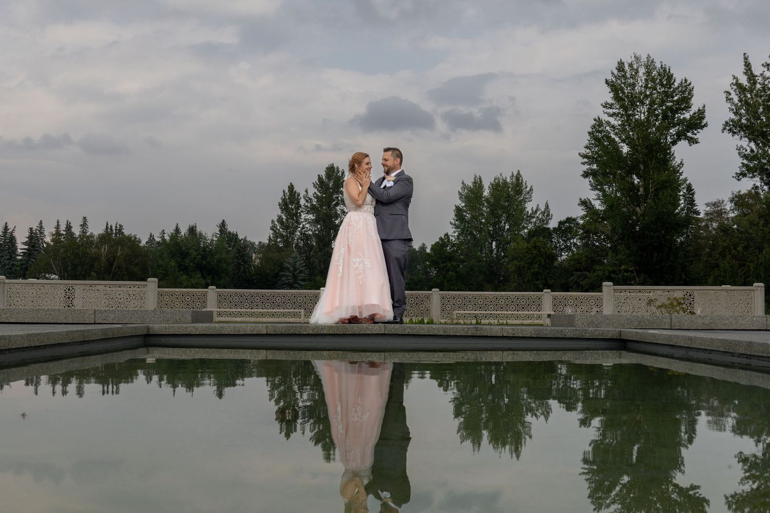 Wedding couple embraces near a reflective pool at dusk with trees in background.