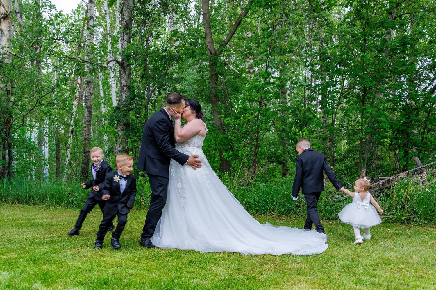 Bride and groom kiss in forest while children in formal attire play around them on green grass.