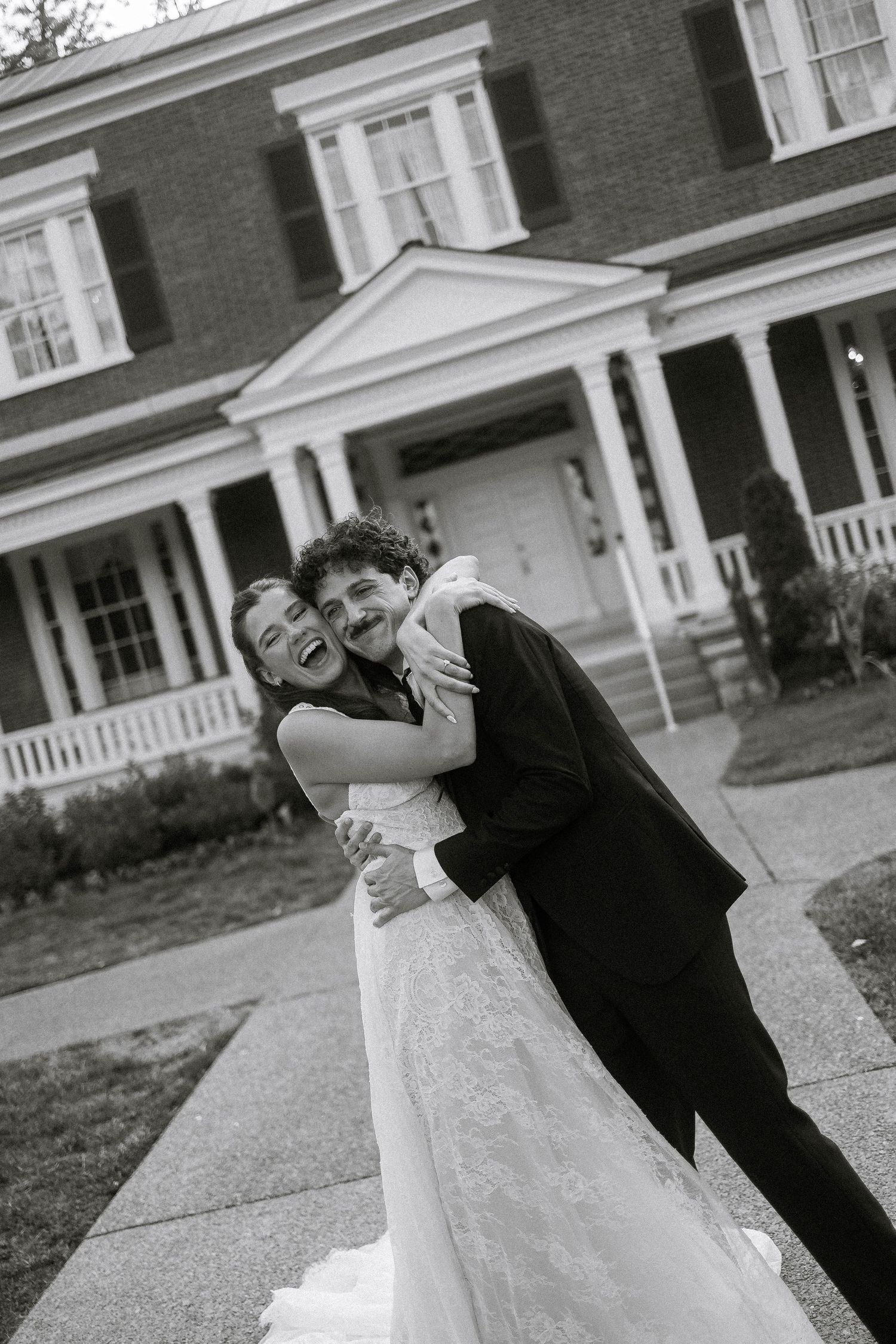Black and white photo of a joyful bride and groom embracing and laughing outside an elegant brick mansion.