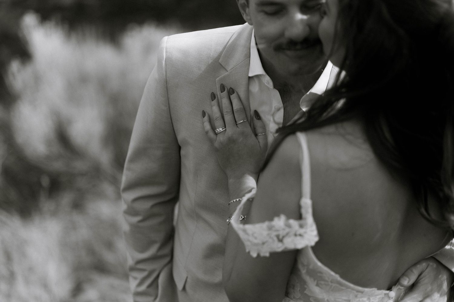 Black and white photo of a couple in wedding attire, bride showing off her ring while groom smiles tenderly.