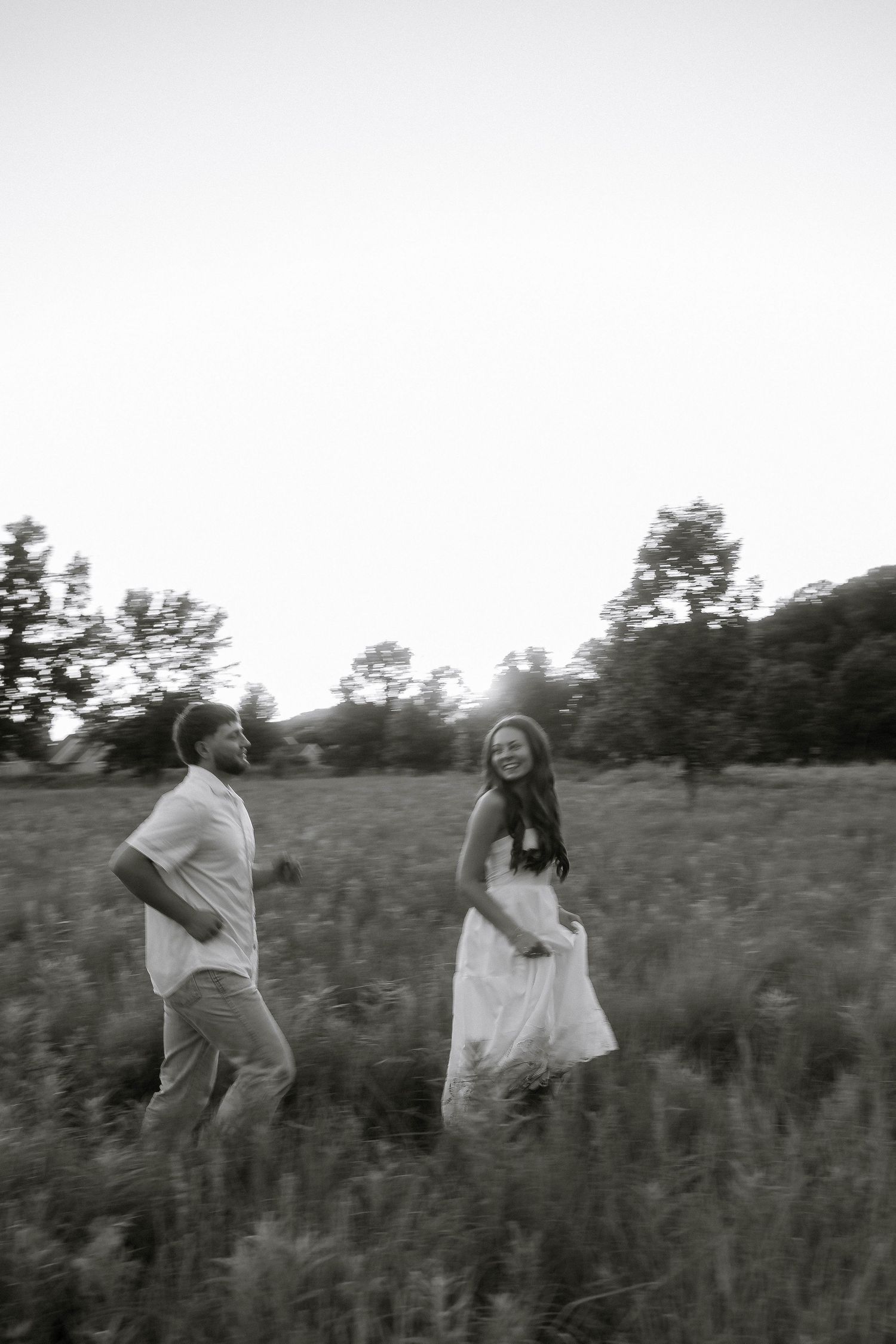 Black and white photo of a couple walking through a field, woman in white dress smiling at man during golden hour.