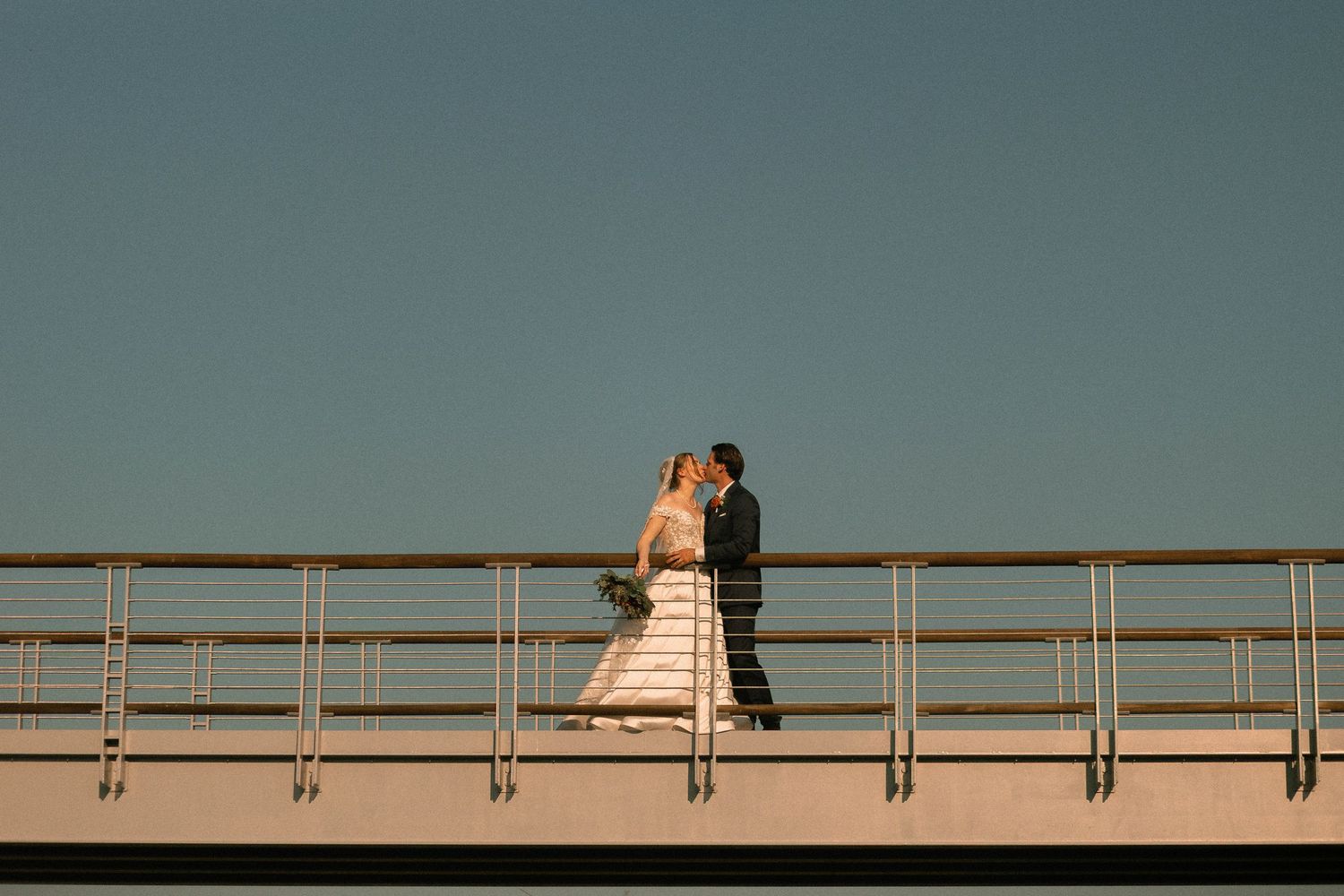 A couple in wedding attire share an intimate moment on a bridge railing against a teal sky.