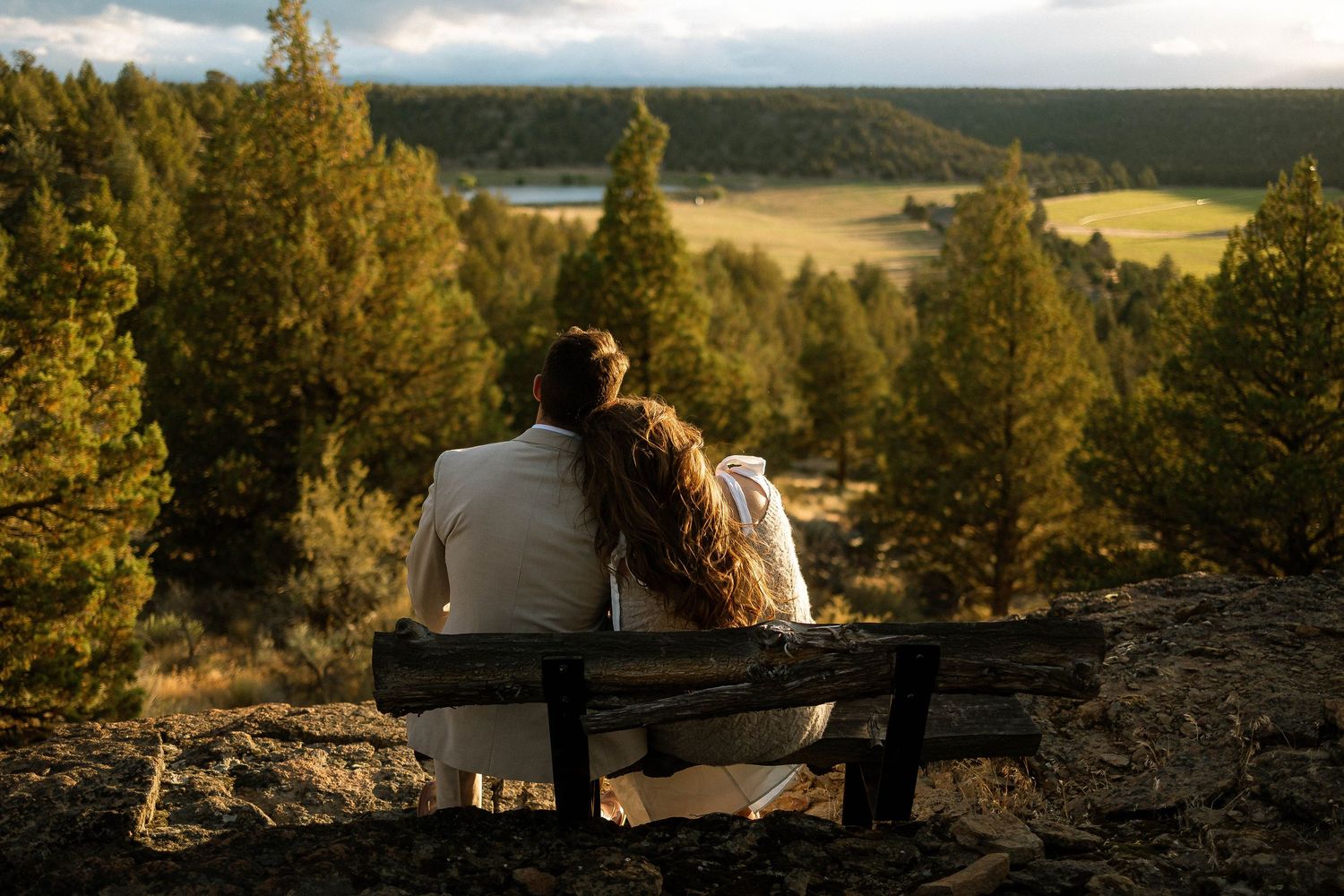 Couple sitting on a bench overlooking a scenic forest valley at sunset in a romantic outdoor setting.