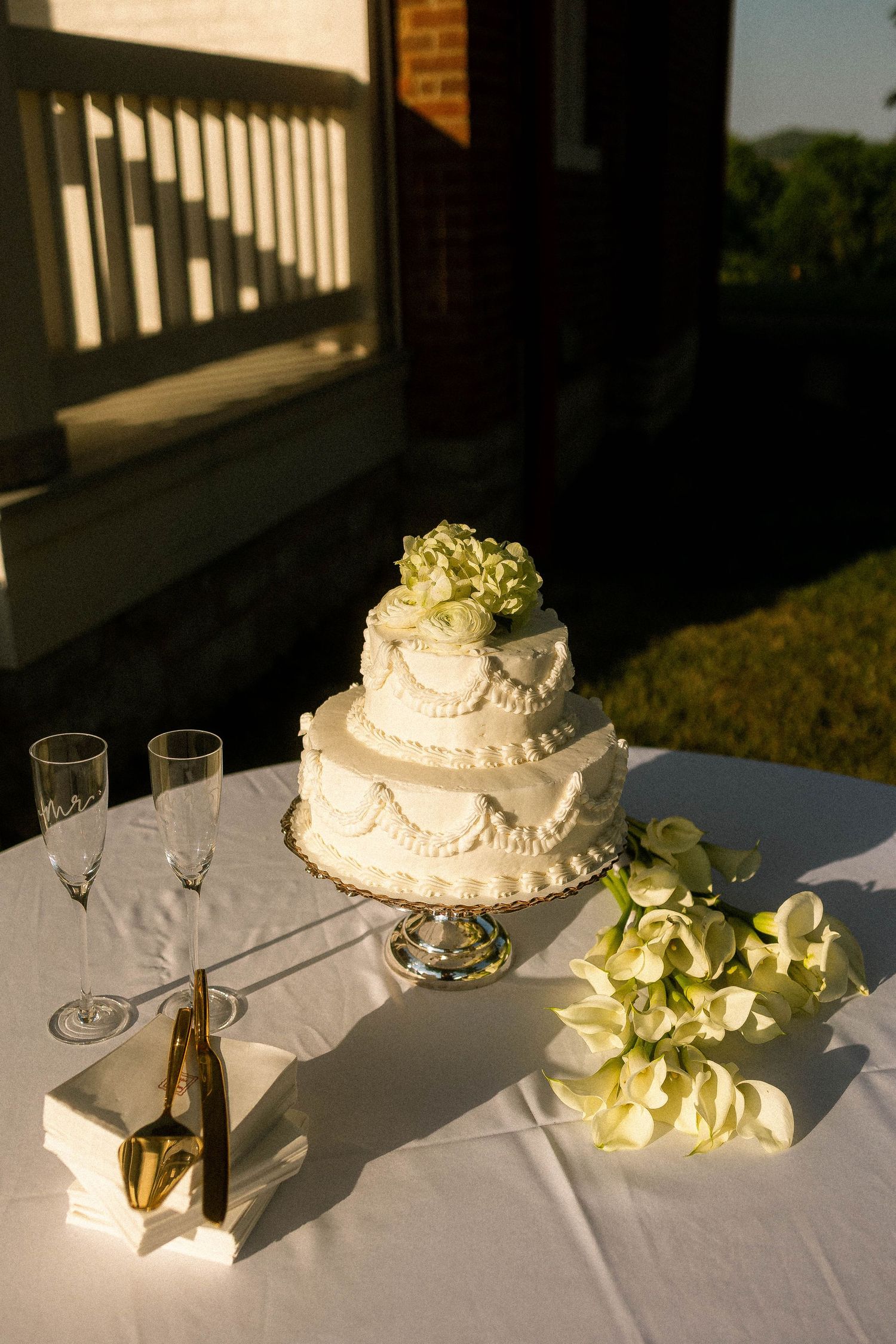 Elegant three-tier white wedding cake with floral topper on silver stand, displayed outdoors with champagne flutes and calla lilies.