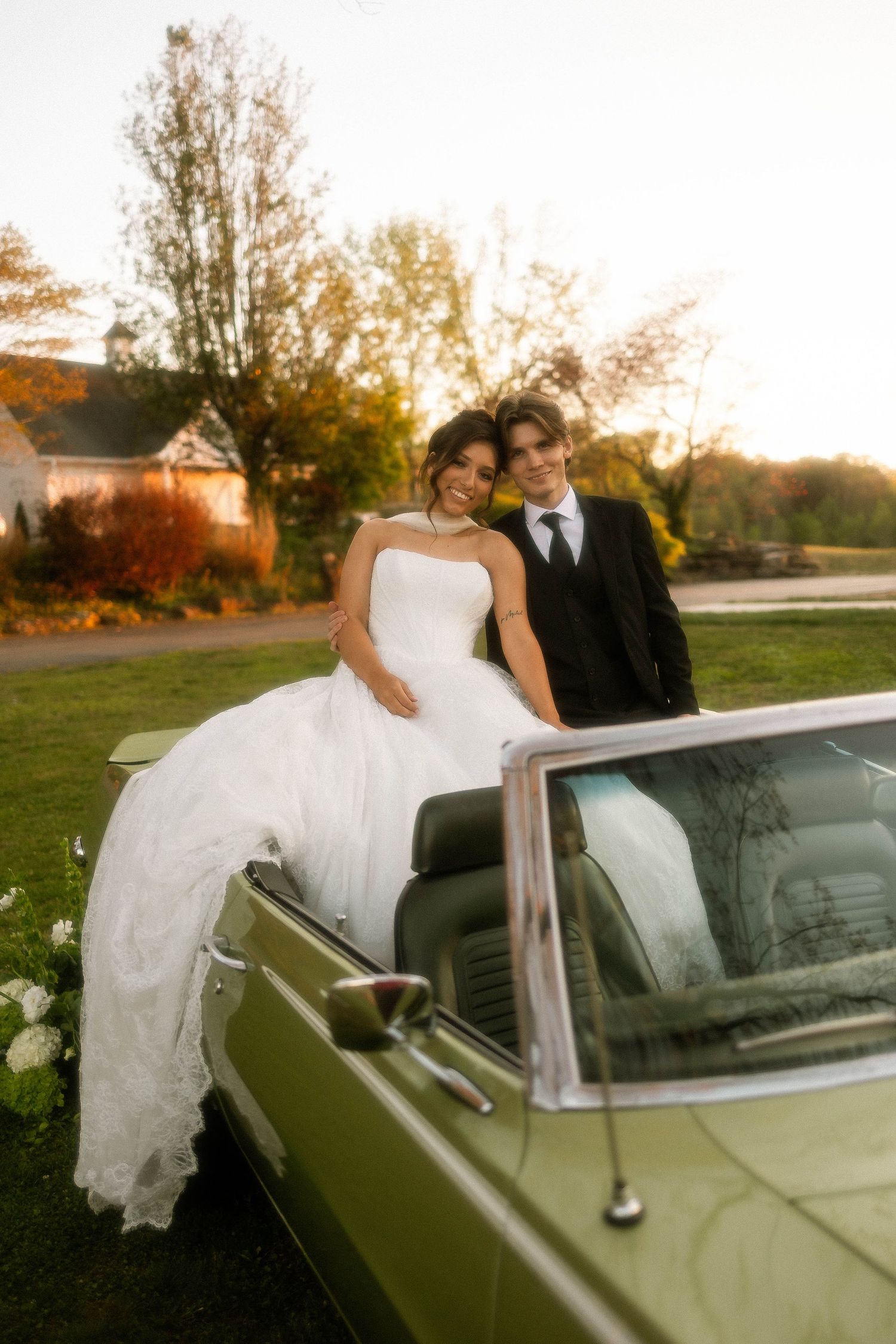 Bride in white gown and groom in black suit pose beside a vintage green convertible at golden hour.