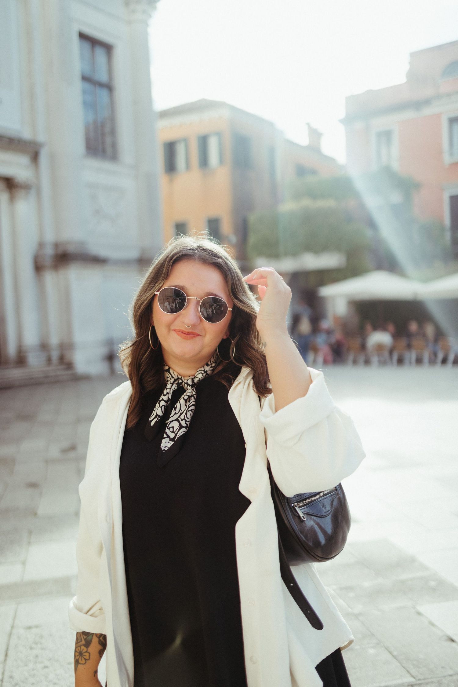 A person wearing sunglasses and a black and white outfit poses outdoors against an urban backdrop.