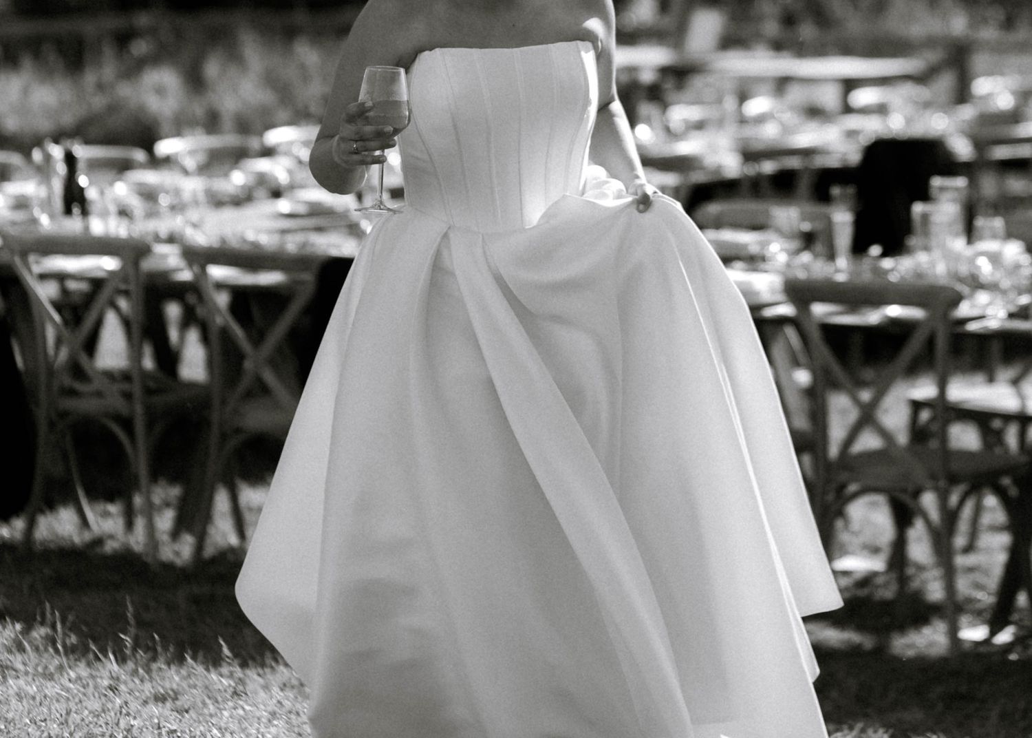 Bride in a strapless white ball gown holding a glass at an outdoor wedding reception in black and white.