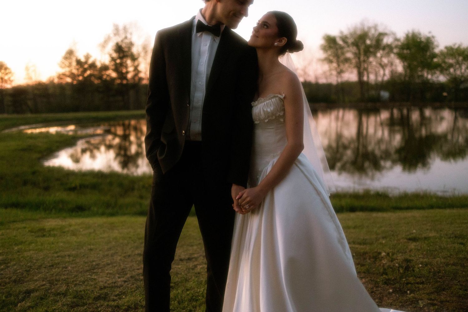 Bride and groom holding hands at sunset near a reflective pond, groom in tuxedo and bride in strapless white gown.