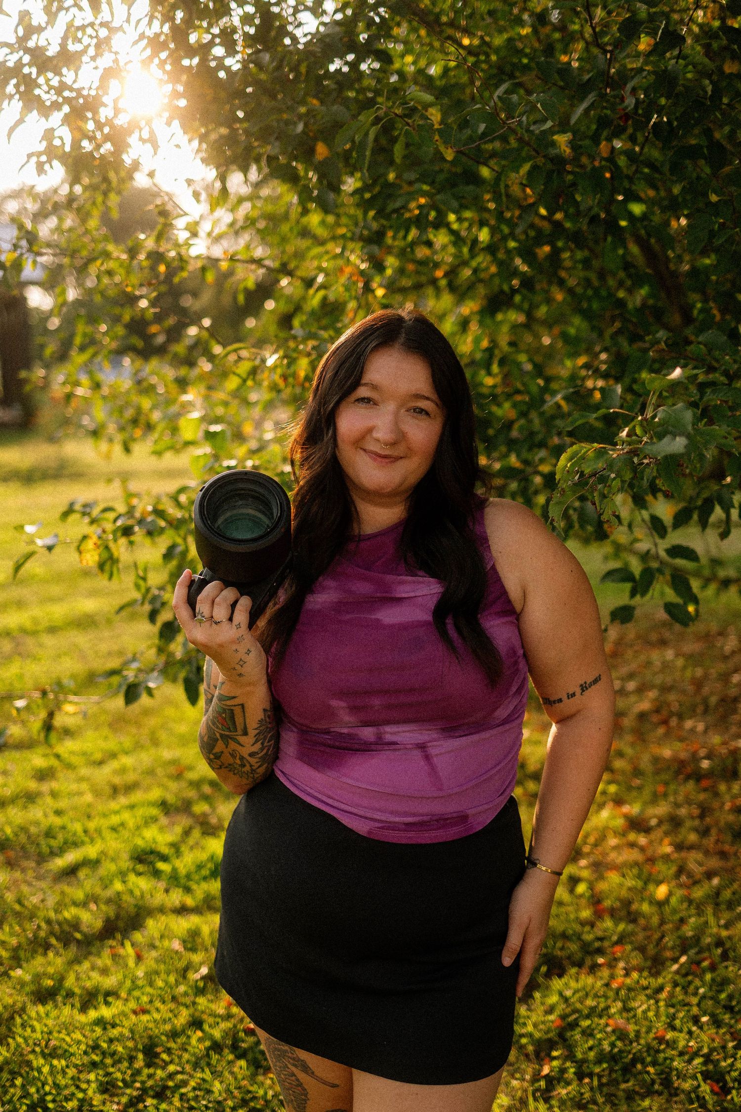 Woman in purple top holding a camera outdoors at golden hour with sunlit green trees in background.