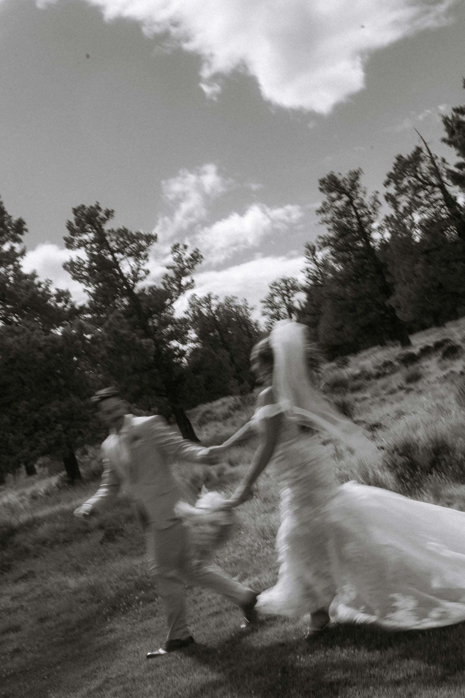 Black and white photo of a bride and groom dancing joyfully outdoors, motion blur capturing their spinning movement among trees.