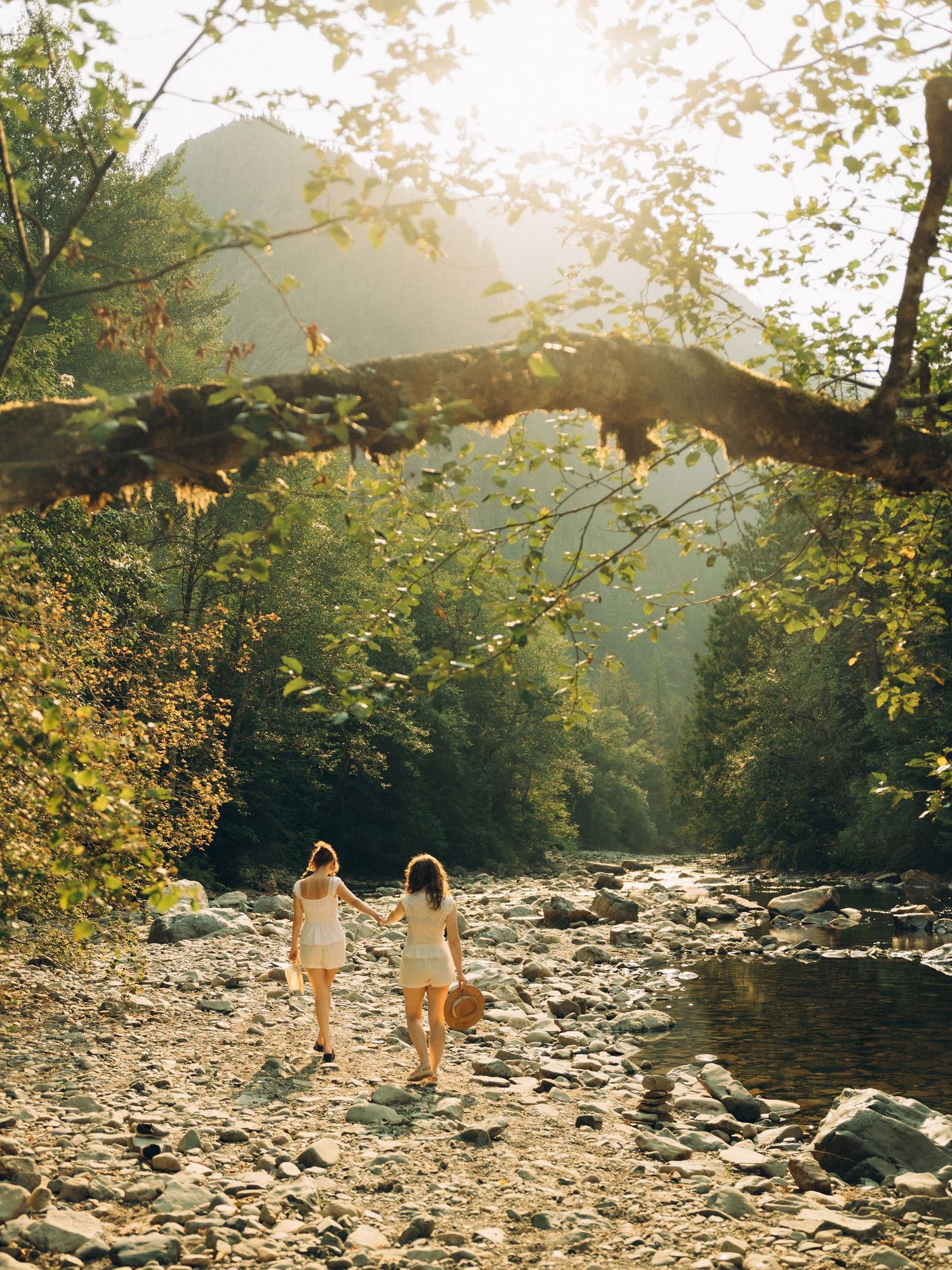 Two women holding hands walk along a rocky riverbank surrounded by lush green trees in golden sunlight.