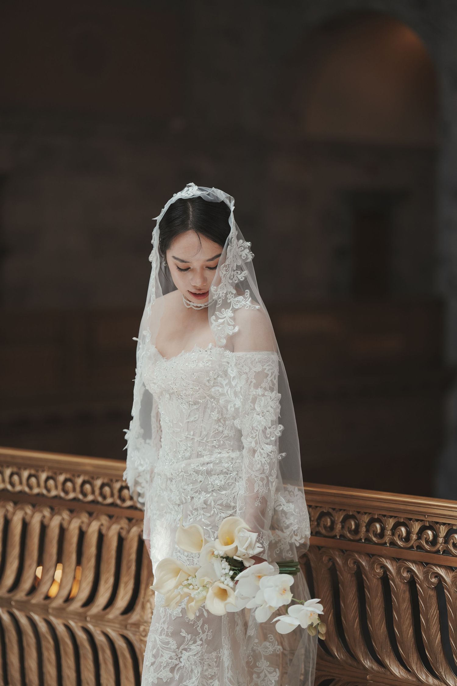 Elegant bride in lace off-shoulder gown and floral veil holds white calla lily bouquet inside ornate venue.